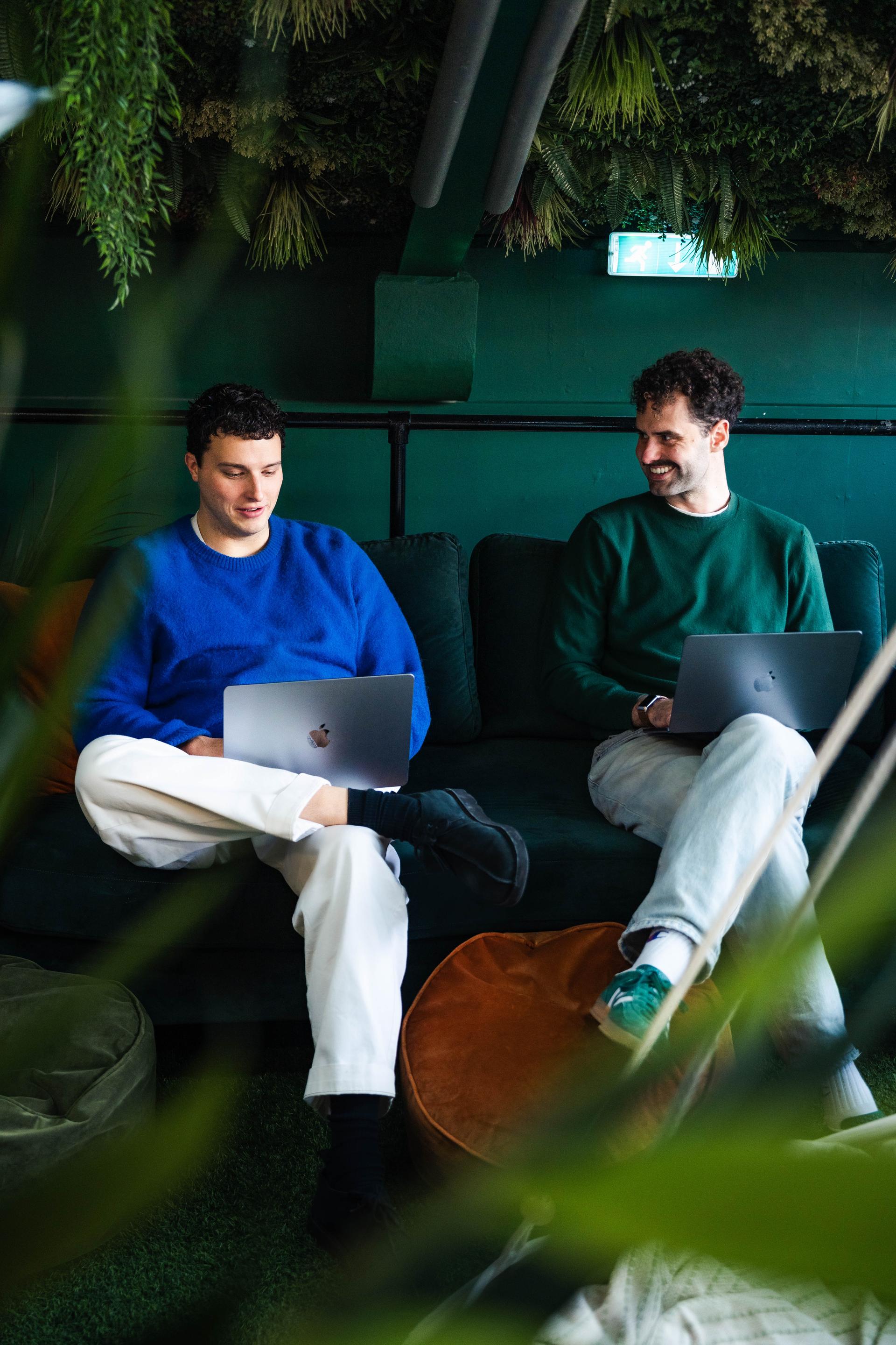 Two young men, one wearing a blue sweater and white pants, the other in a green sweater and jeans, sit on a green couch with laptops, surrounded by lush indoor plants in a cozy workspace.