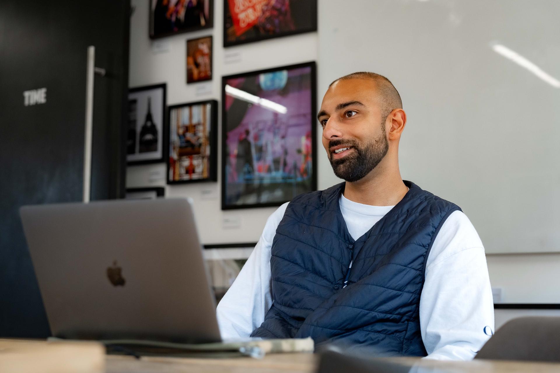 A bearded man in a navy vest and white shirt sits at a wooden desk with a laptop, smiling while talking. Behind him, a whiteboard and framed pictures decorate the modern office space.