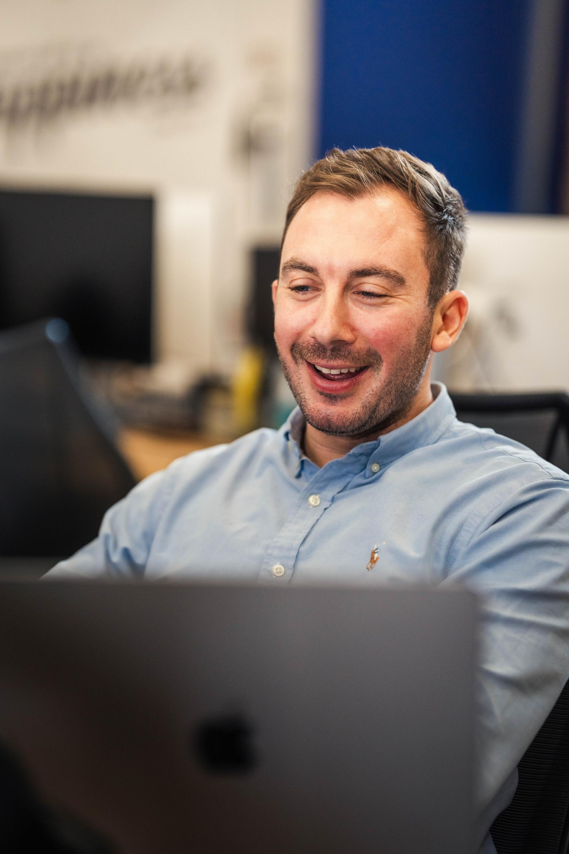 A cheerful man wearing a light blue Ralph Lauren shirt is focused on his laptop in a modern office setting, surrounded by blurred computer monitors and a motivational wall sign that reads "Happiness" in the background.