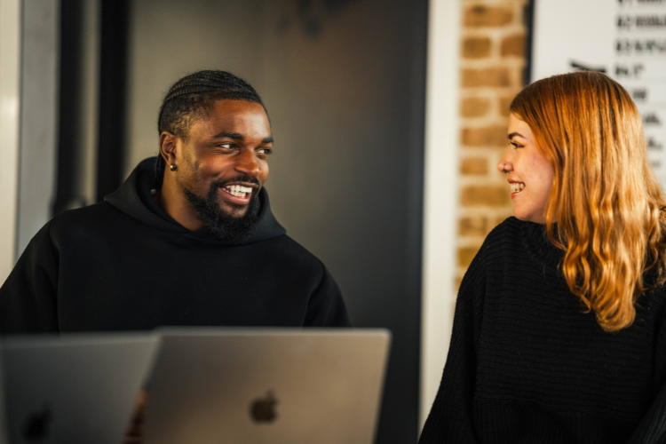 A man and a woman in black sweaters sit at a table with laptops, smiling warmly at each other in a modern office setting.