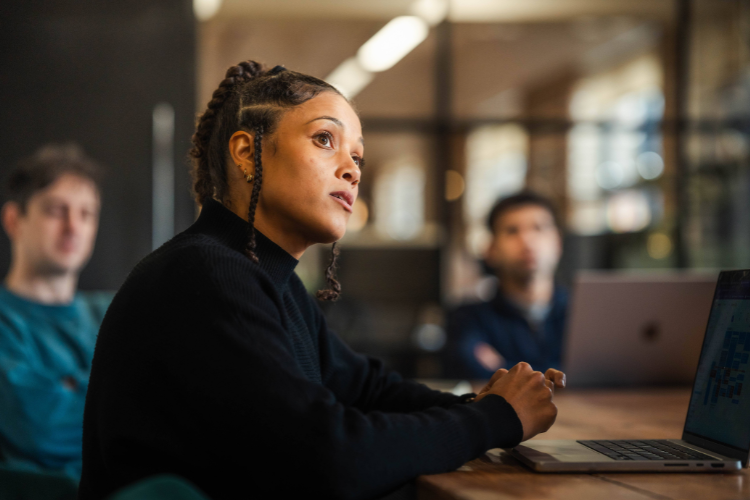 A serious woman in a black turtleneck, with braids, listens attentively during a discussion, with blurred teammates in the background.