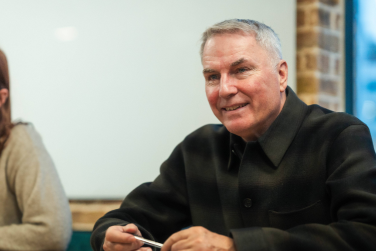A senior man in a dark plaid coat listens attentively in a modern office with a whiteboard behind him.
