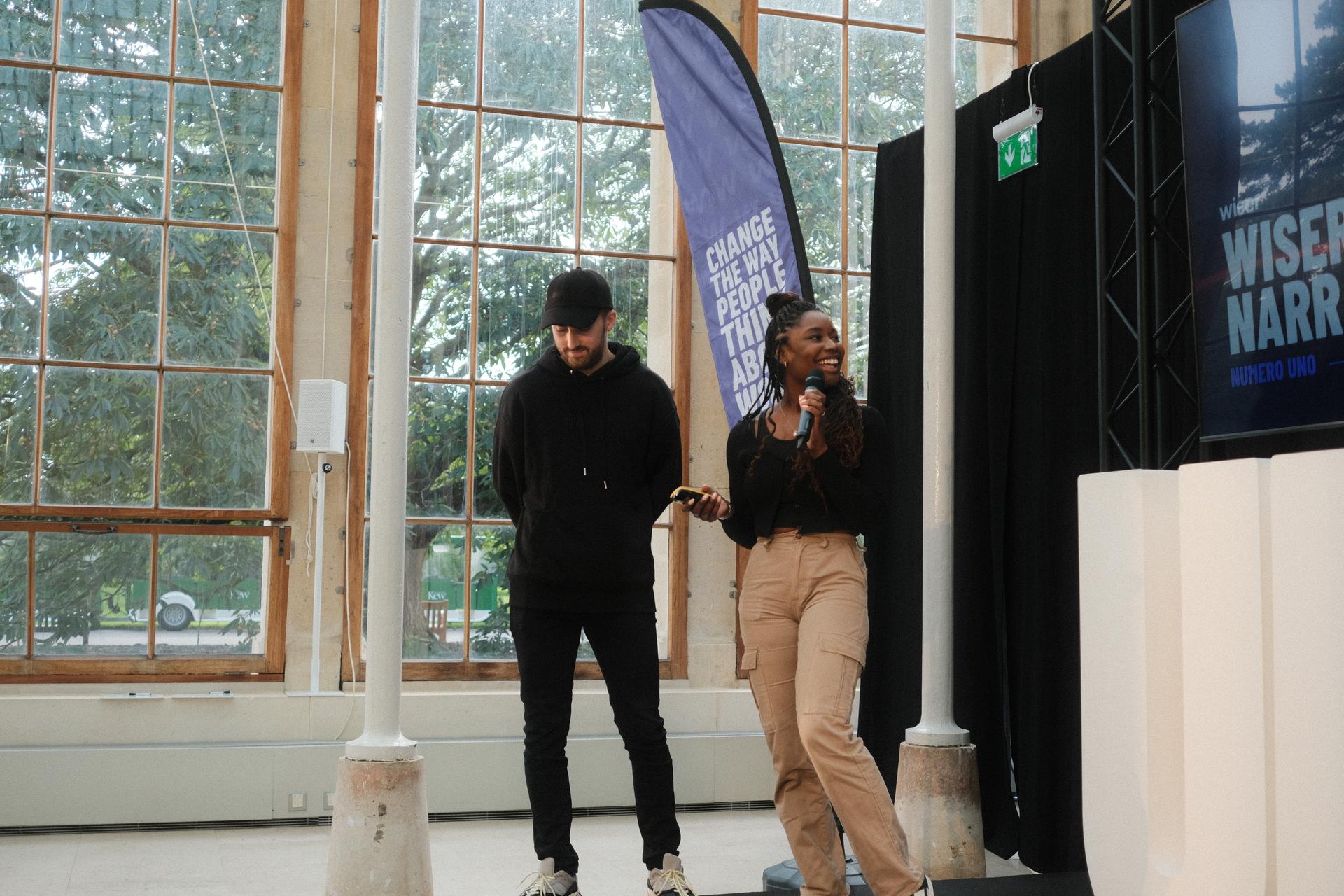 A woman in cargo pants speaks into a microphone while smiling, alongside a man in black attire on stage. Behind them, a banner and a large screen display event-related text in a bright venue.