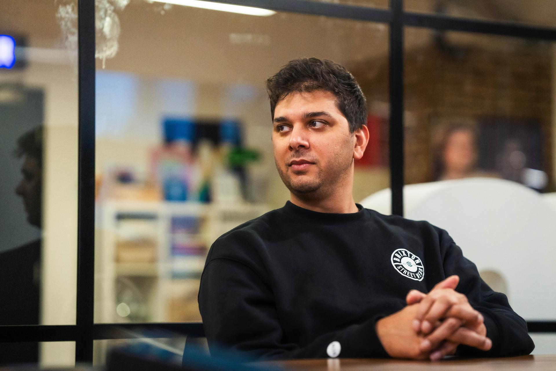 A young man in a black sweatshirt with a circular logo sits at a table in a contemporary office setting, hands clasped, gazing to the side with a thoughtful expression.