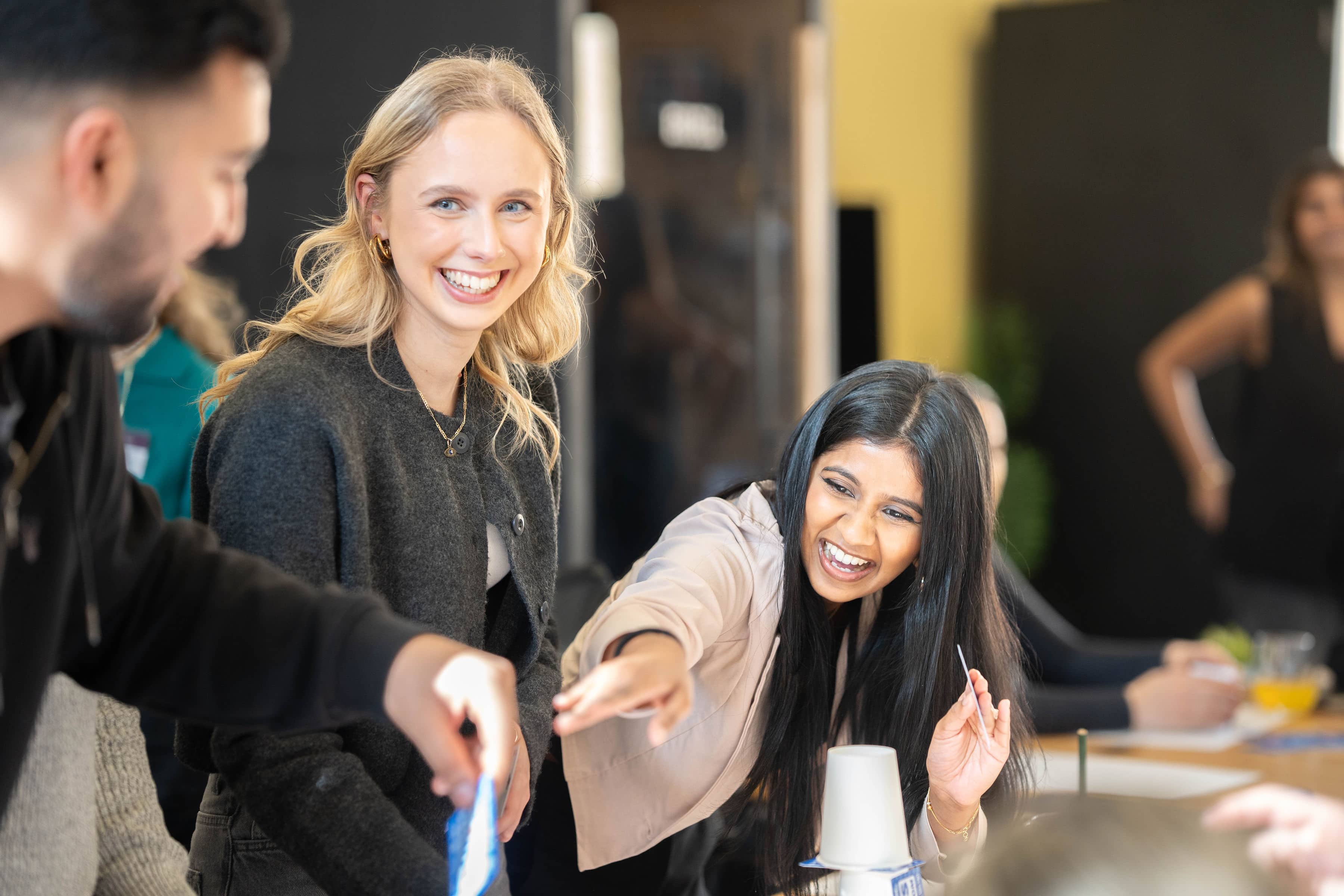 Two women laugh and interact during a group activity, pointing and engaging with others around a table.