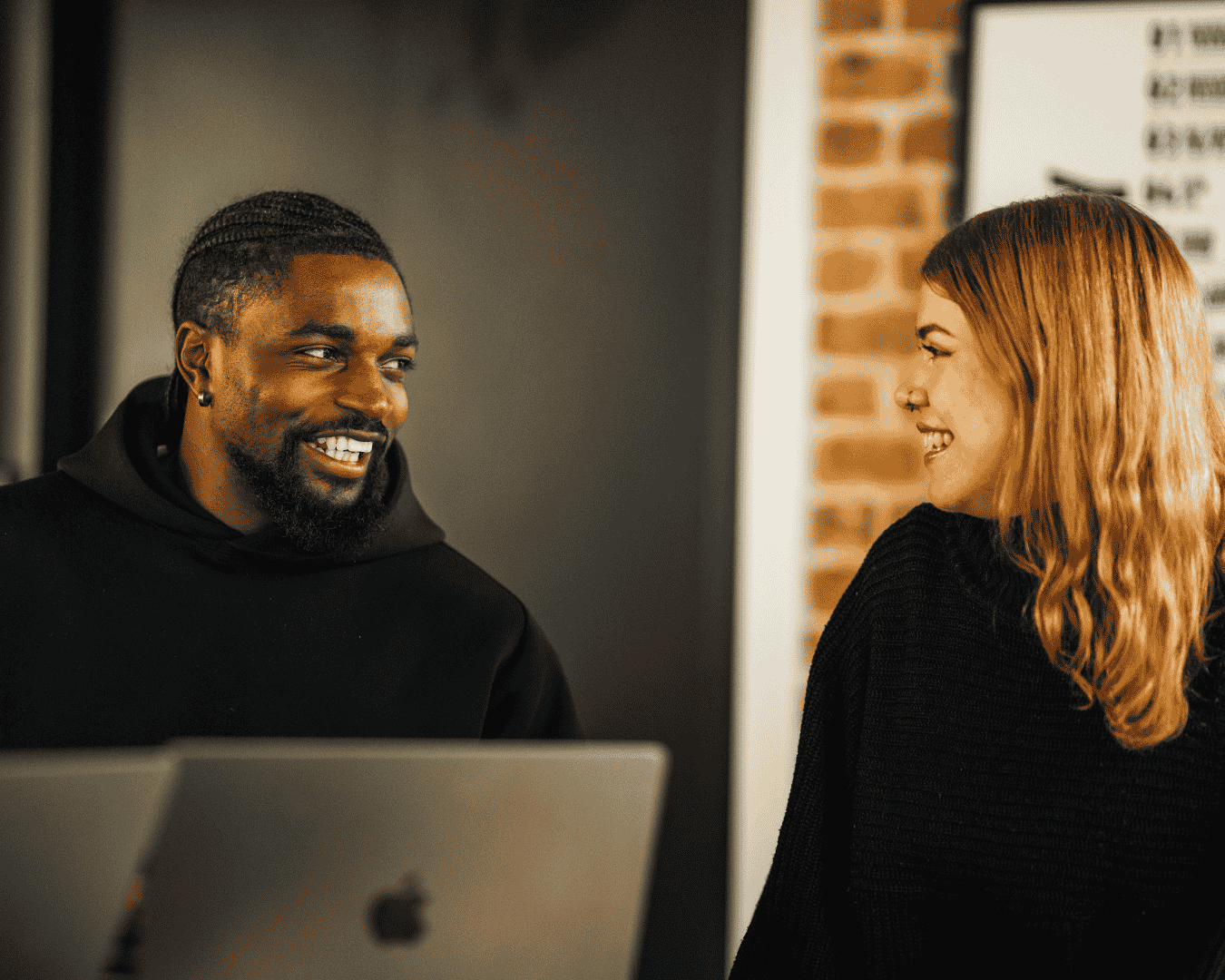 A man and a woman in black sweaters sit at a table with laptops, smiling warmly at each other in a modern office setting.