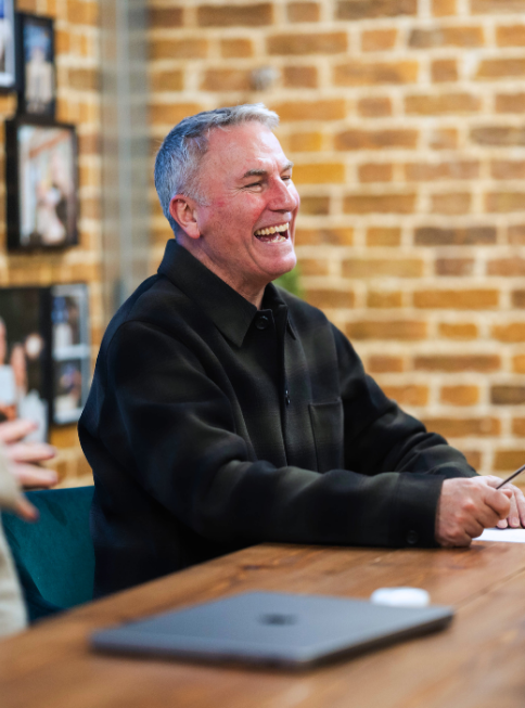 A cheerful older man in a black jacket laughs while seated at a wooden table, with a laptop in front.