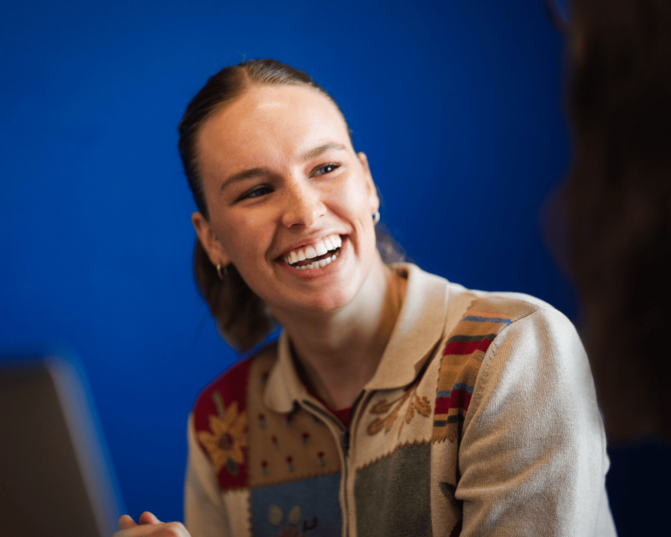 Smiling woman in a patterned cardigan engaged in conversation, seated indoors against a vibrant blue background.