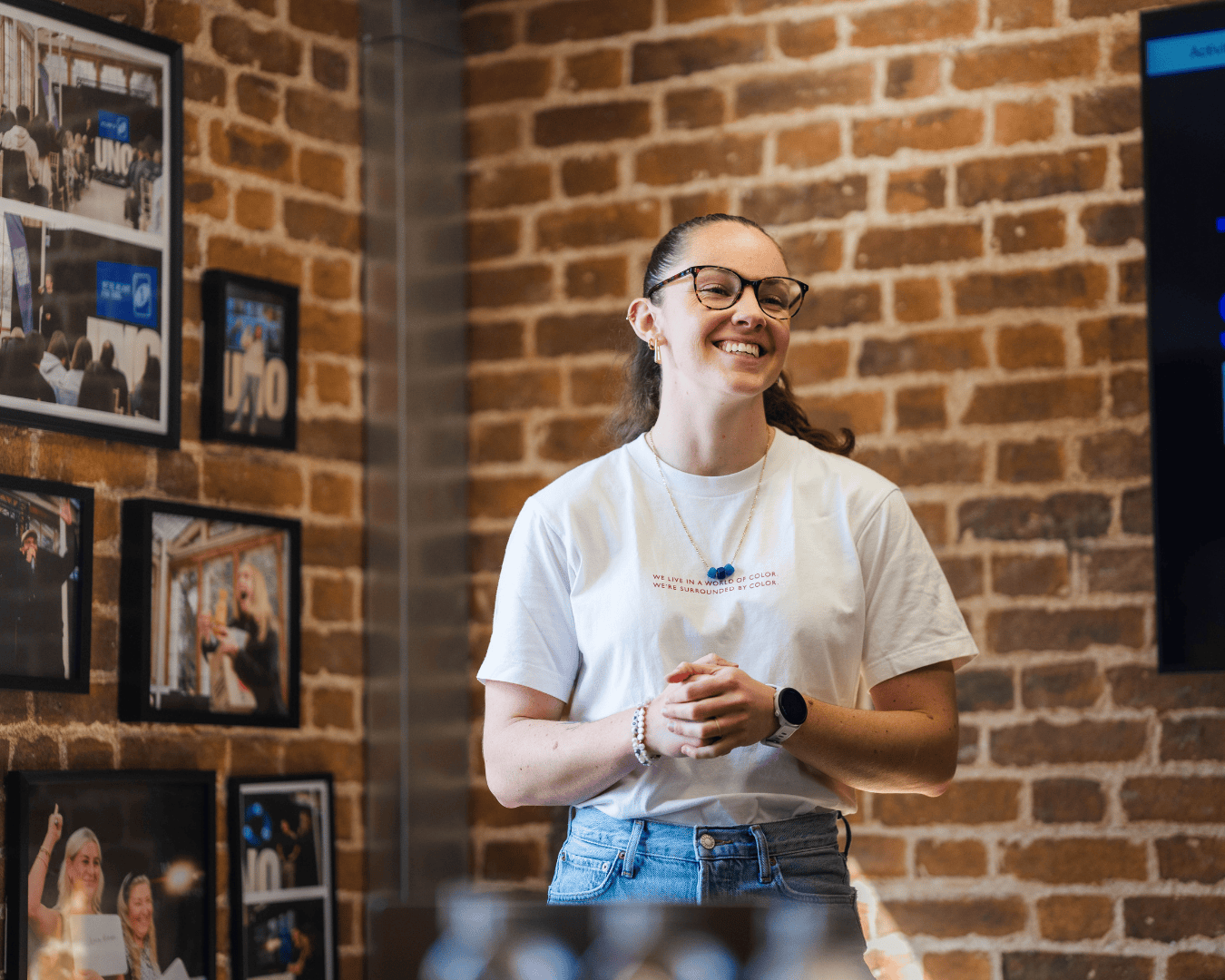 Smiling woman giving a presentation in a modern office with brick walls, wearing a white t-shirt and glasses, standing near framed photos and a screen.