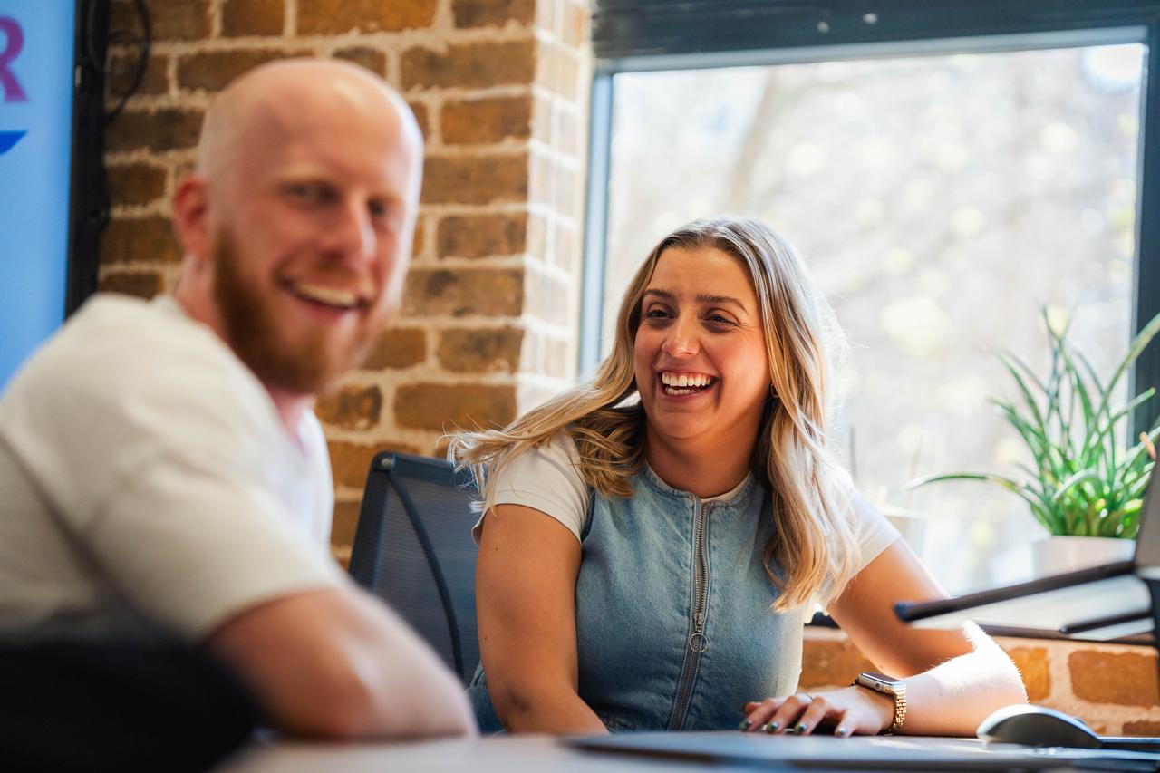 A smiling man and woman enjoy a lighthearted conversation while seated at a desk in a sunlit office, featuring exposed brick walls, a large window, and a green potted plant.