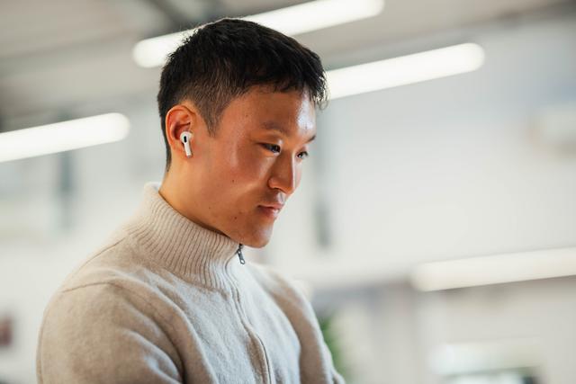 A young man with a calm expression, dressed in a beige sweater and wearing AirPods, concentrates intently in a modern, well-lit office environment with blurred lights in the background.