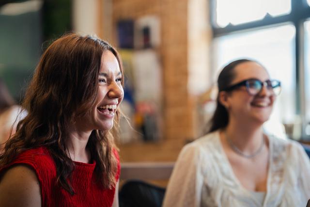 Two women, one in a red top and the other in a white blouse with glasses, laugh joyfully while sitting in a warmly lit, casual office environment near a window.