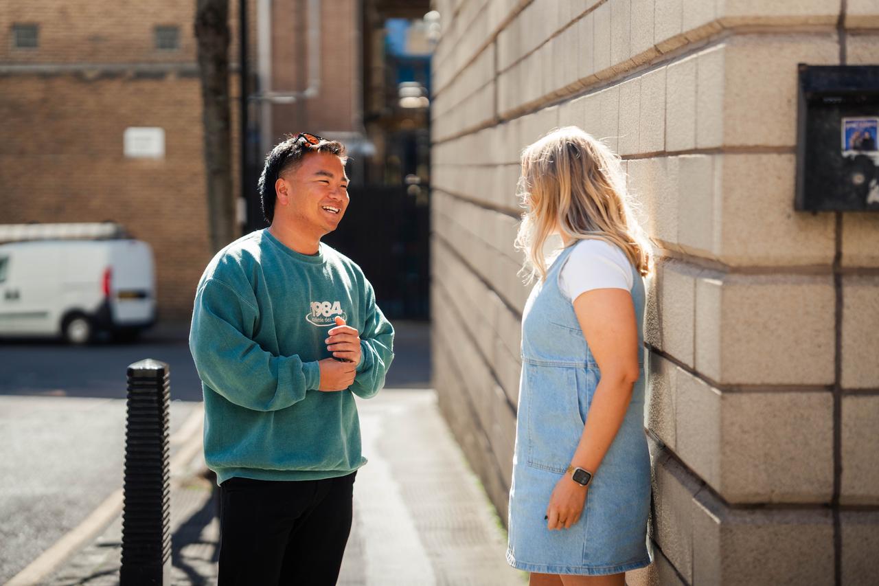 A casually dressed man and woman enjoy a sunny conversation outside. The woman leans against a brick wall while the man, smiling, stands facing her with a van parked nearby.