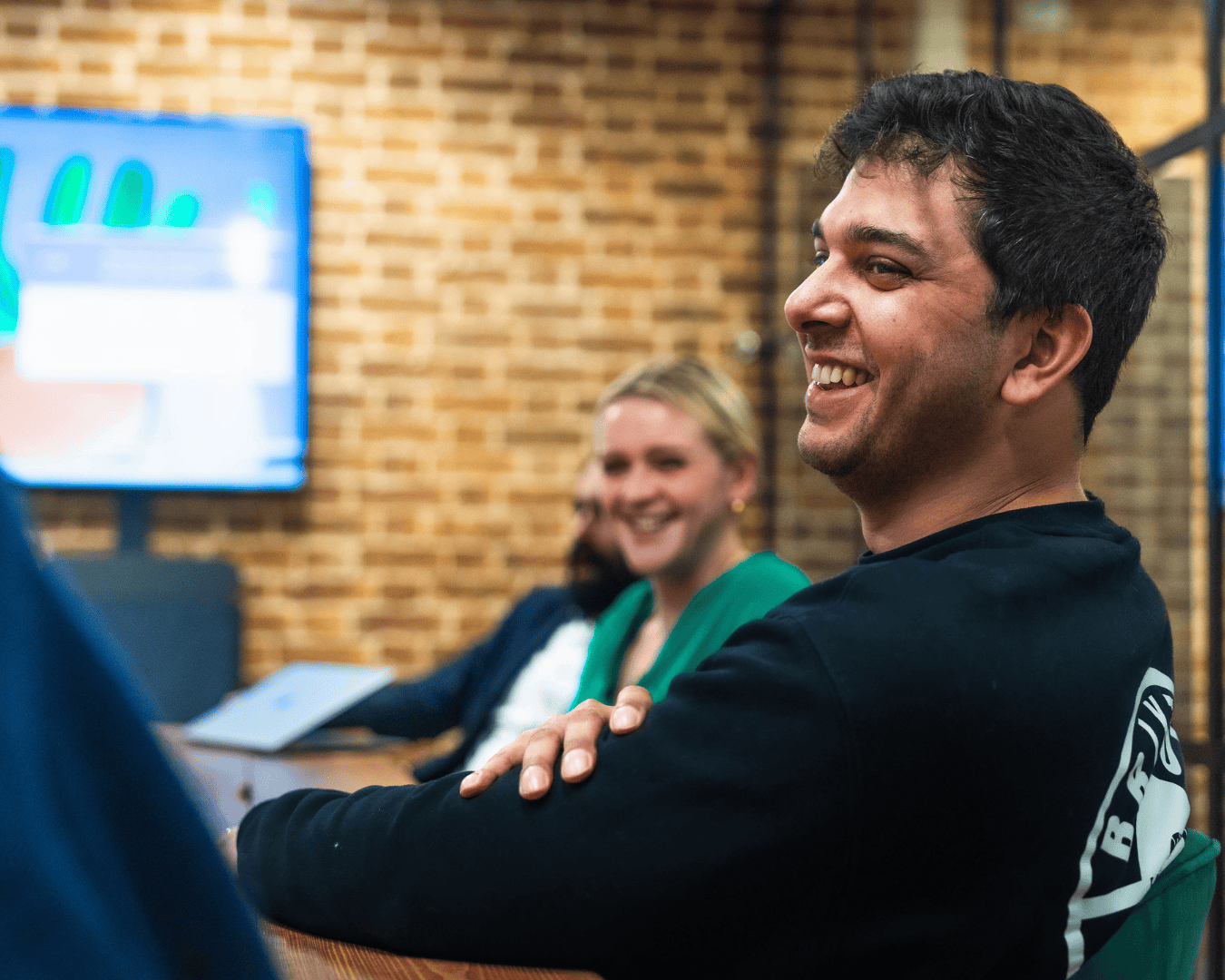 A smiling man sits at a wooden table in a modern office, engaged in conversation with colleagues. A woman in green and a man in the background also appear cheerful, with a presentation screen and exposed brick wall behind them.