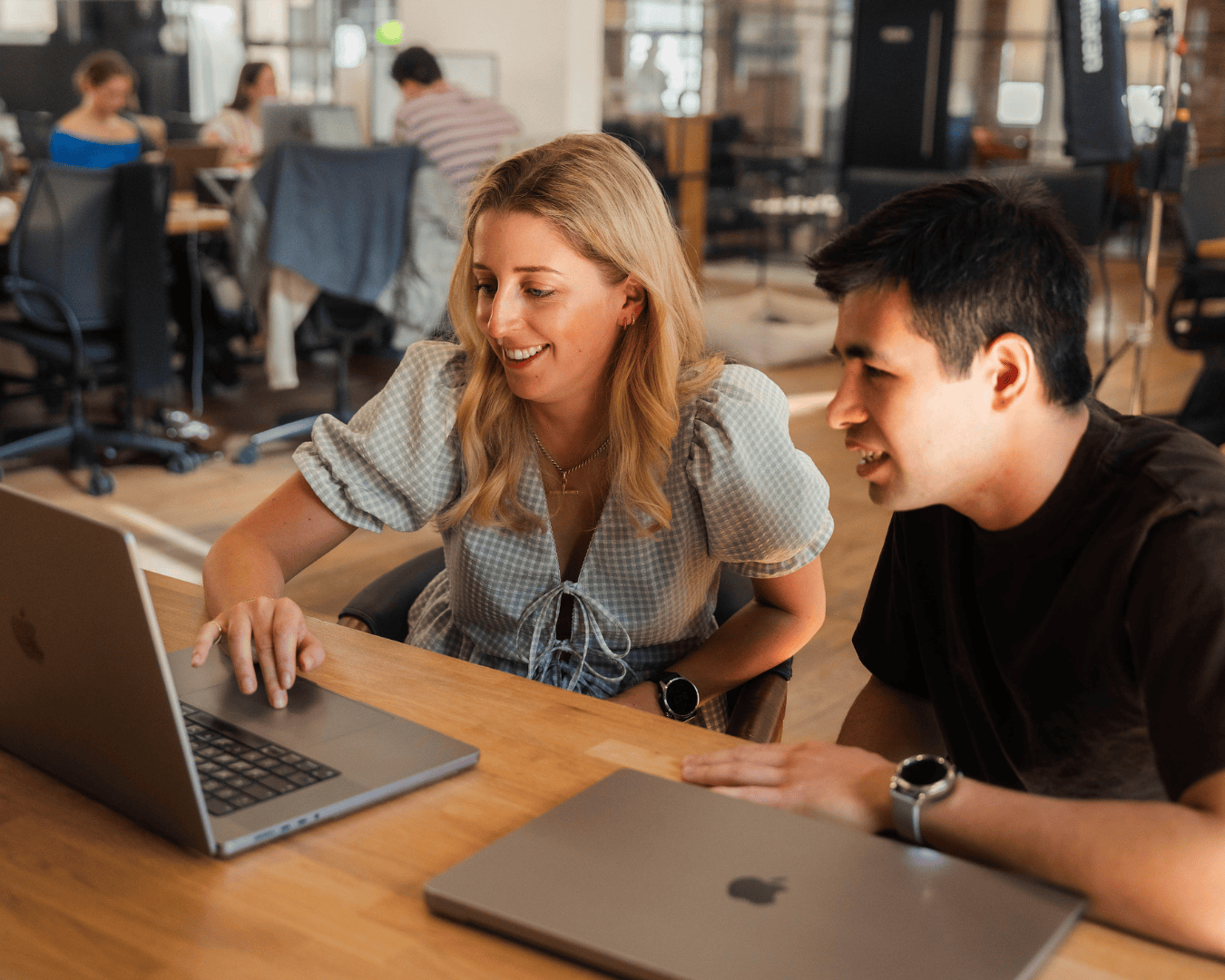 A woman and man sit at a wooden desk in a bright office, smiling as they look at a laptop screen. Other colleagues work in the background of the open-plan space.