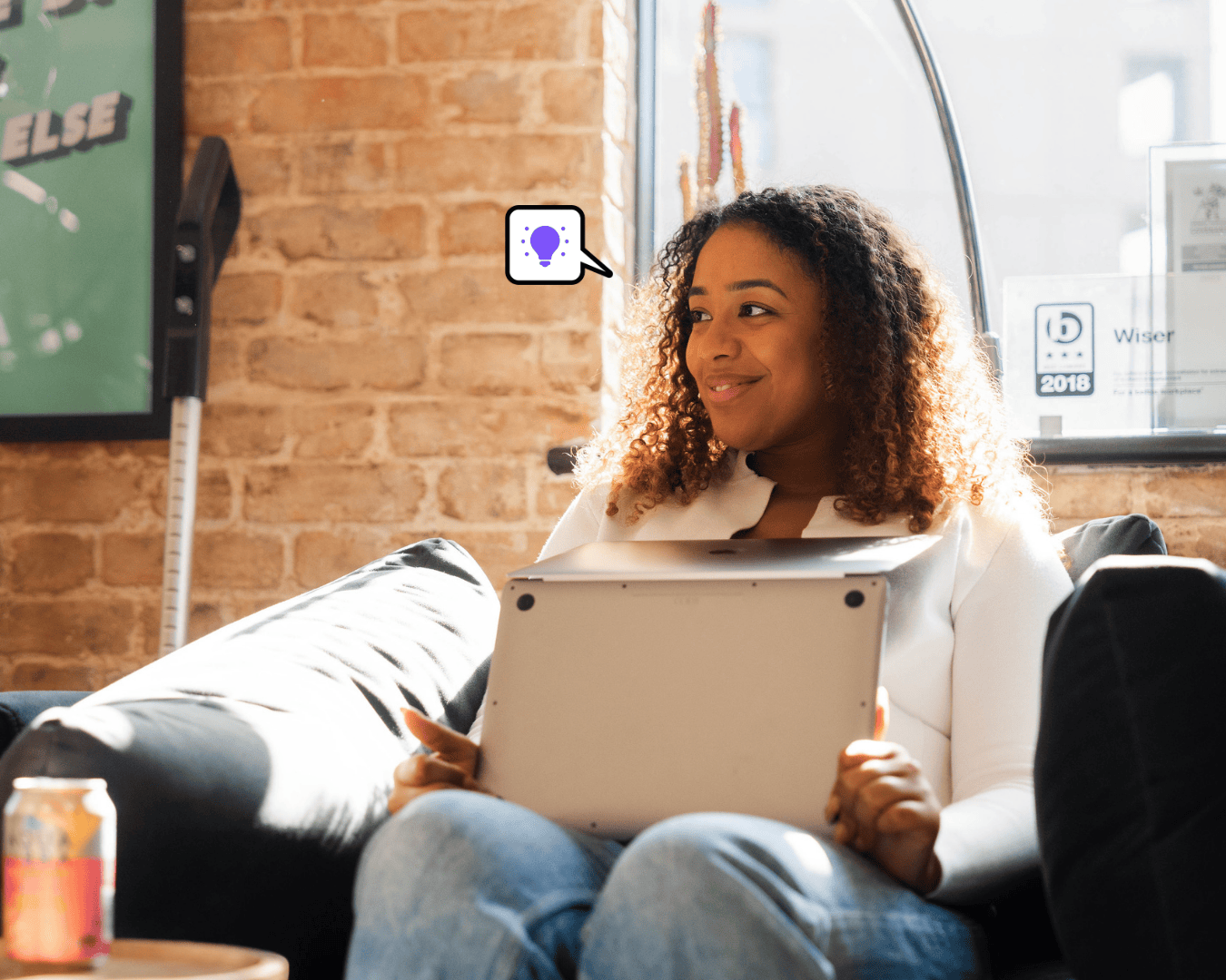 Smiling woman with curly hair sitting on a sofa, holding a laptop, in a bright modern office with exposed brick walls.