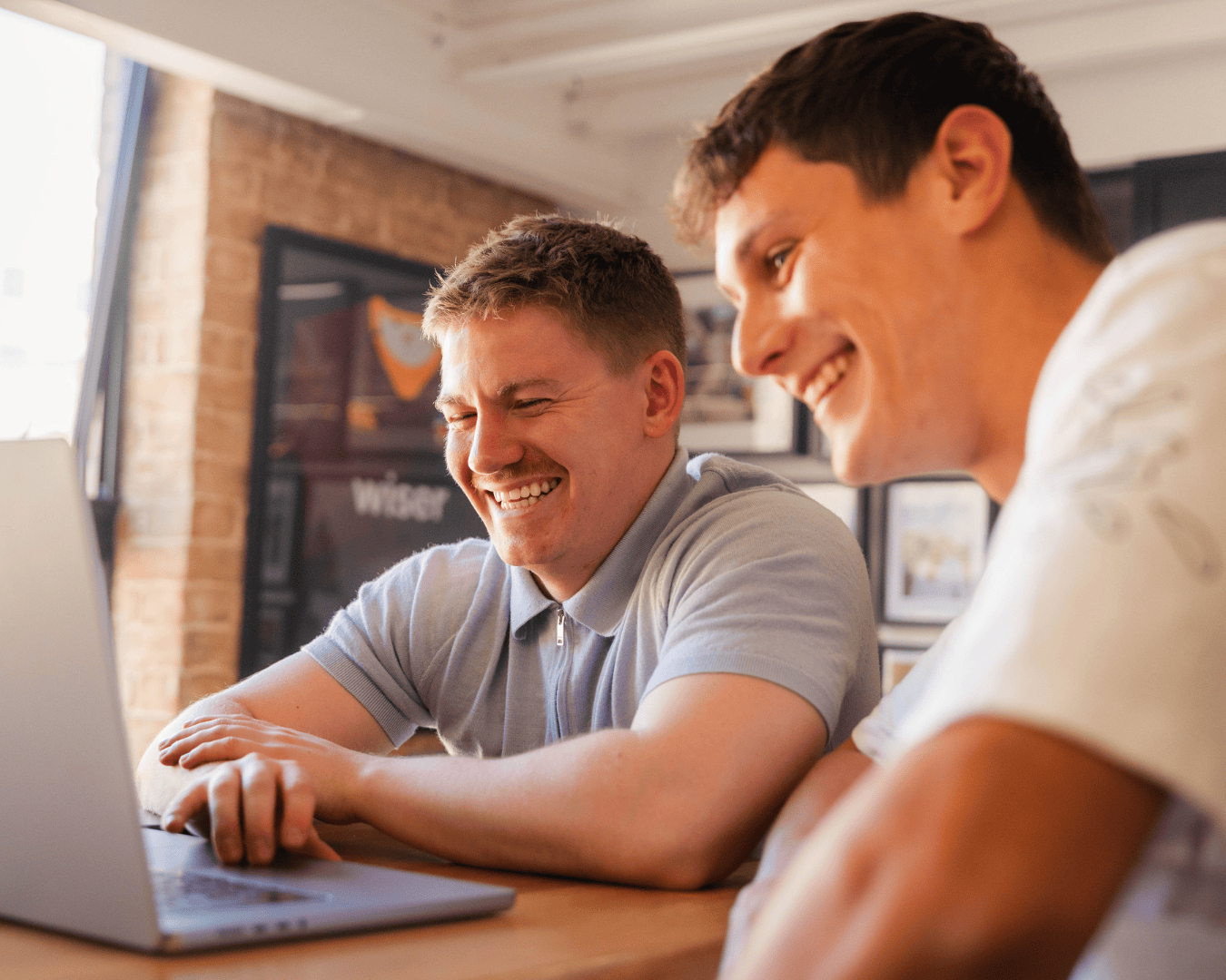 Two men laughing while working together on a laptop in a casual, creative office space with warm lighting.