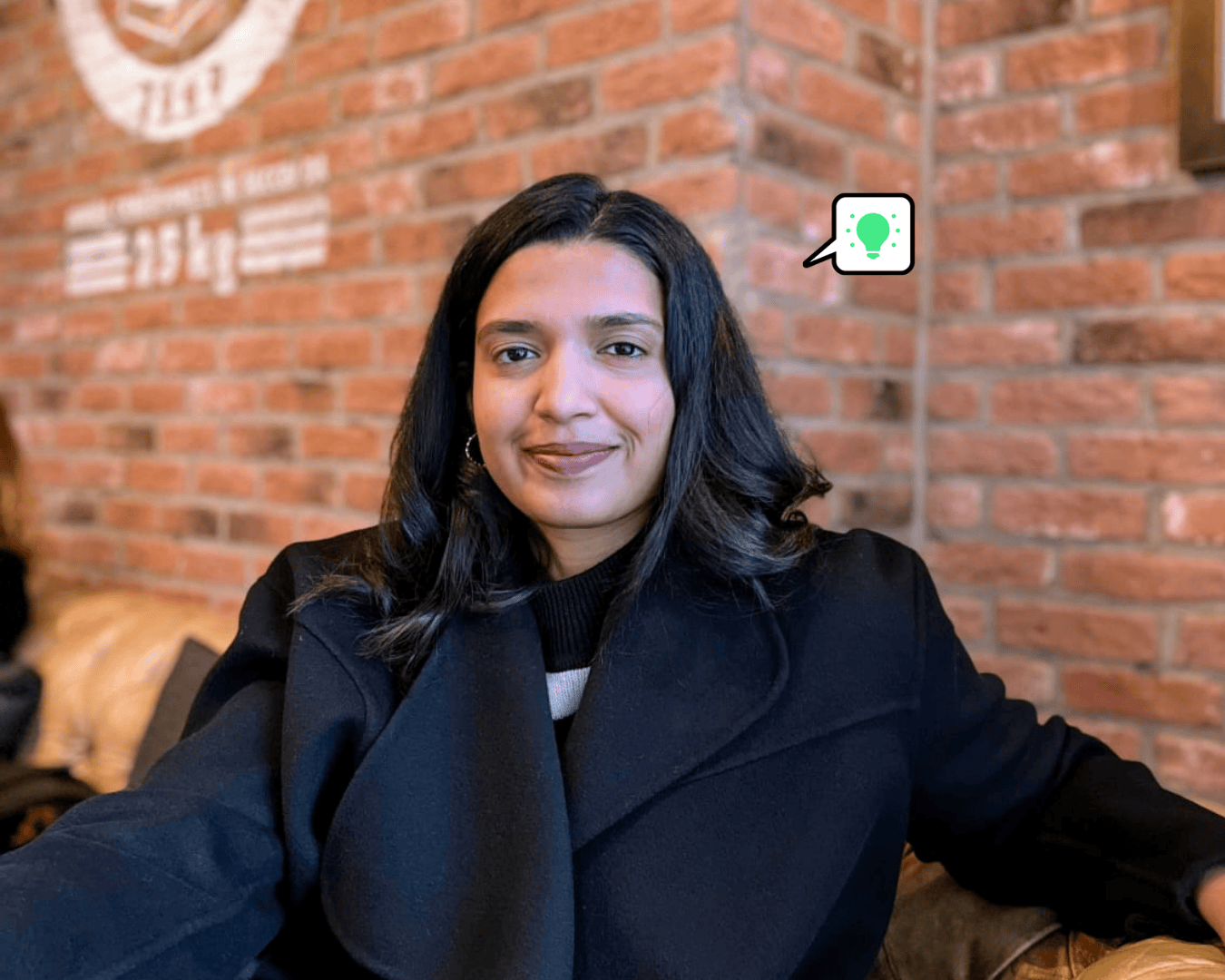 Swapni Mehta, a woman with long dark hair, sits on a leather sofa in a café with brick walls. She is smiling gently and wearing a dark coat.