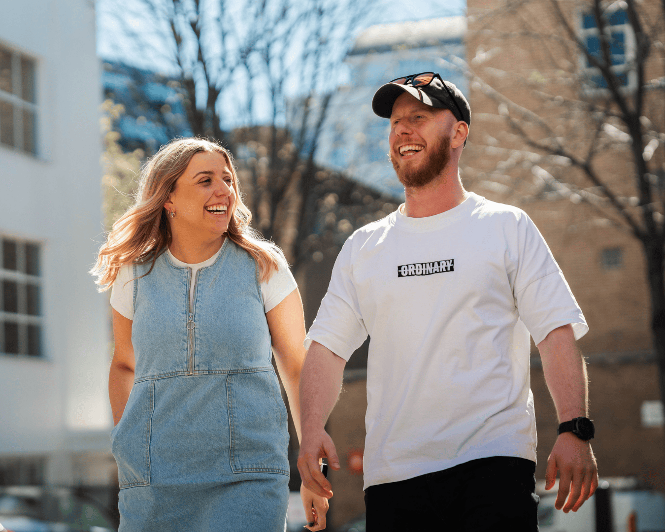 Two people, a woman in a denim dress and a man in a white t-shirt and cap, walk outdoors on a sunny day, smiling and laughing together.