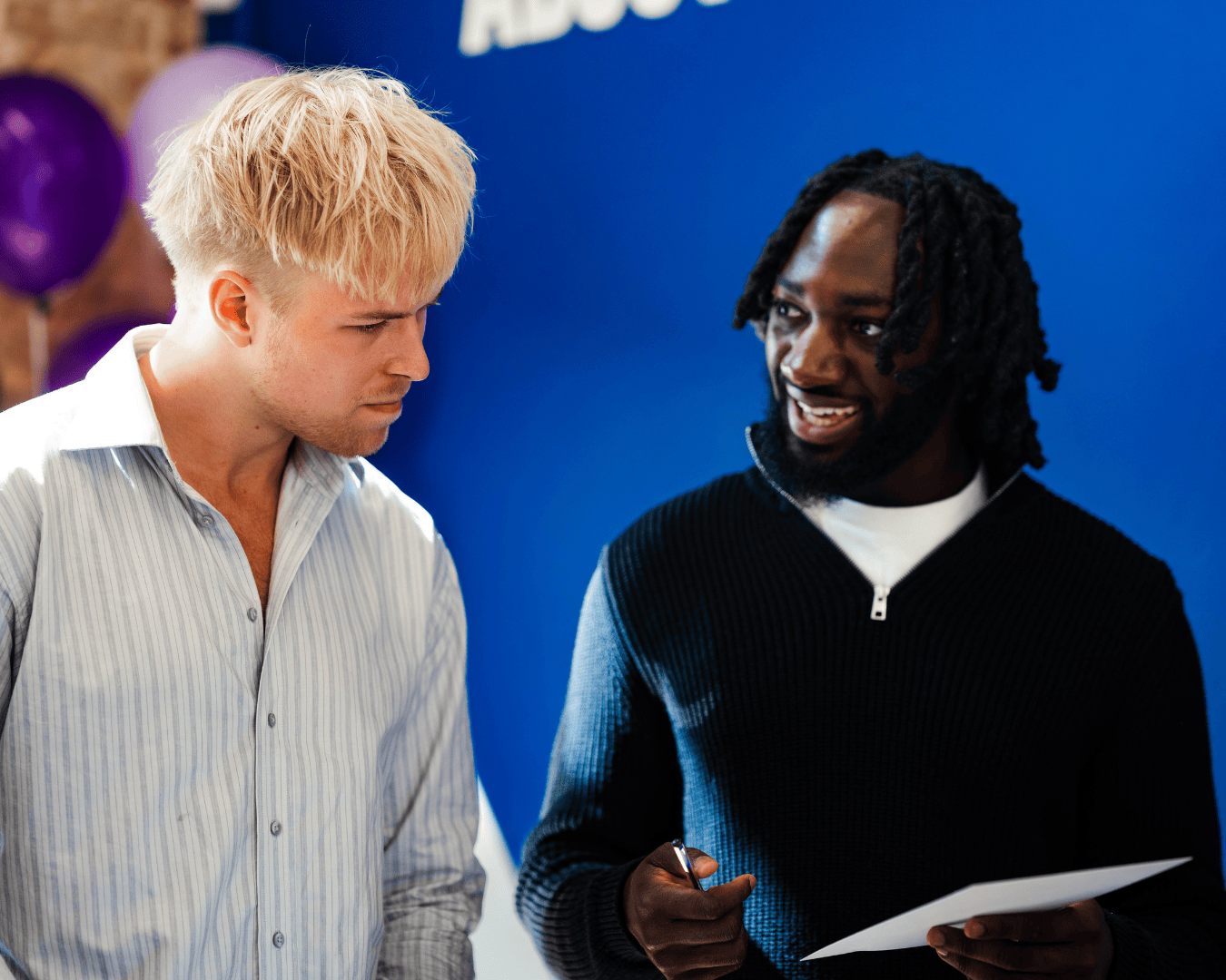 A Wiser Academy ambassador holding a pen and paper smiles while advising a fellow student in a striped shirt at a careers event. Set against a bright blue backdrop with balloons, the image captures an engaging peer-to-peer moment focused on employability support, confidence building, and inclusive career development.