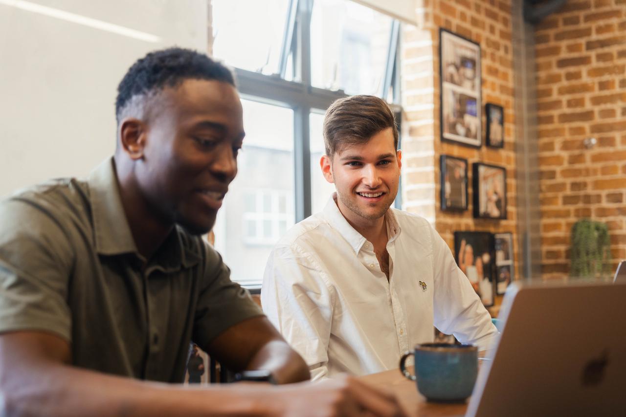 A Black man with short hair and a white man with side-parted hair smile while reviewing digital content. Part of the global employer branding initiative and employee-generated content drive.