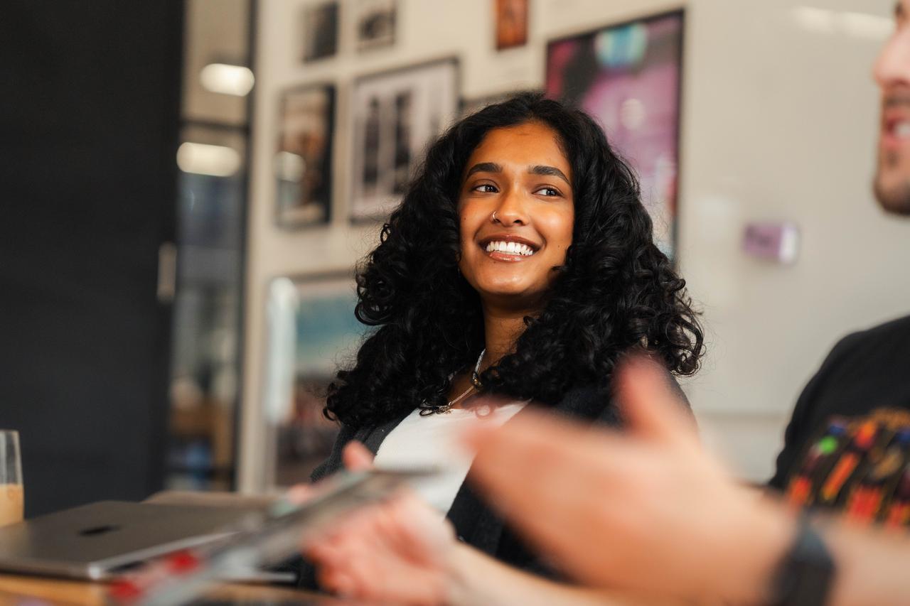 A South Asian woman with long curly hair smiles during discussion. She’s a DNV brand advocate, involved in storytelling workshops that support employee-generated content and social media strategy.
