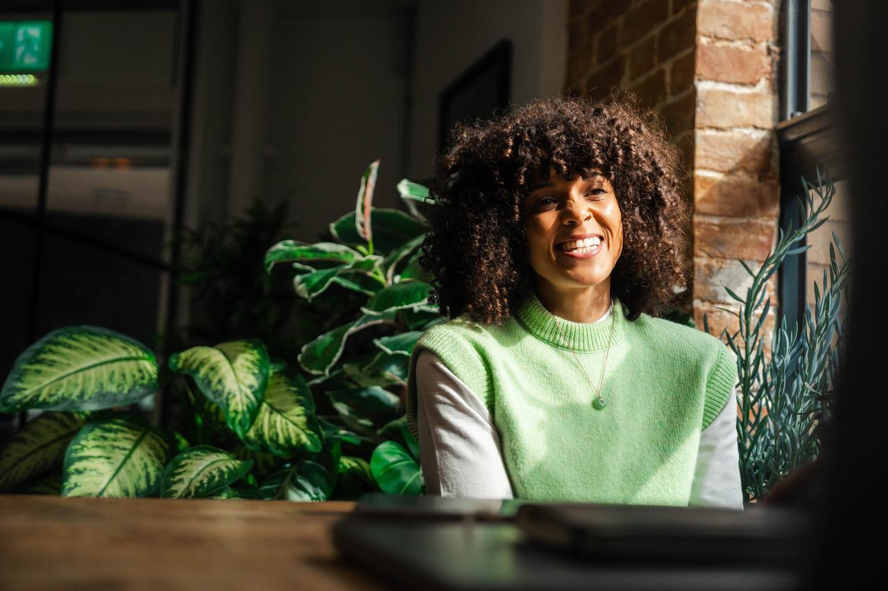 A Black woman with curly hair smiles in sunlight, seated by plants. Part of DNV’s employee advocacy programme, spotlighting EVP activation strategy and employee-generated content in authentic environments.