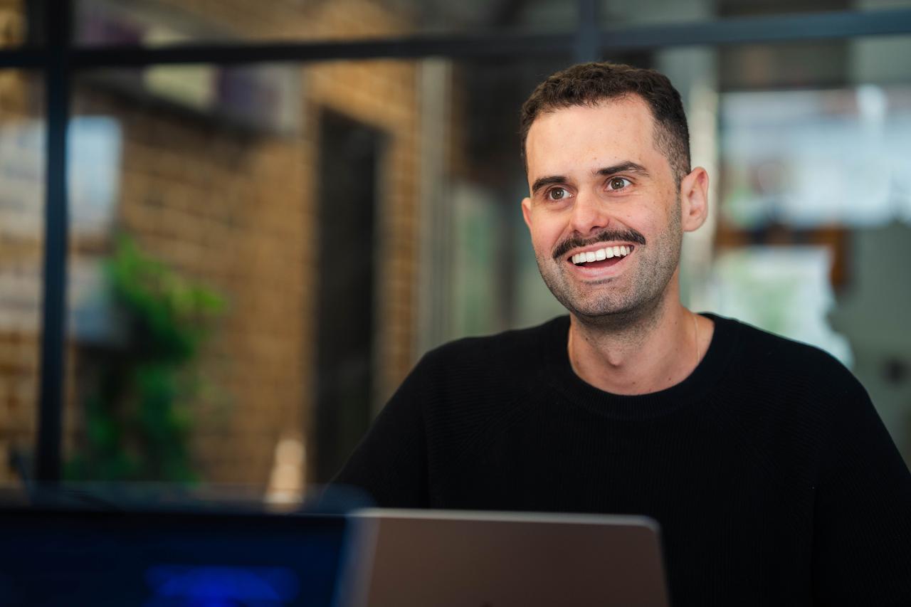 A white man with short dark hair, a moustache and black sweater smiles at a desk with a laptop. He’s participating in a brand storytelling session focused on EVP strategy.