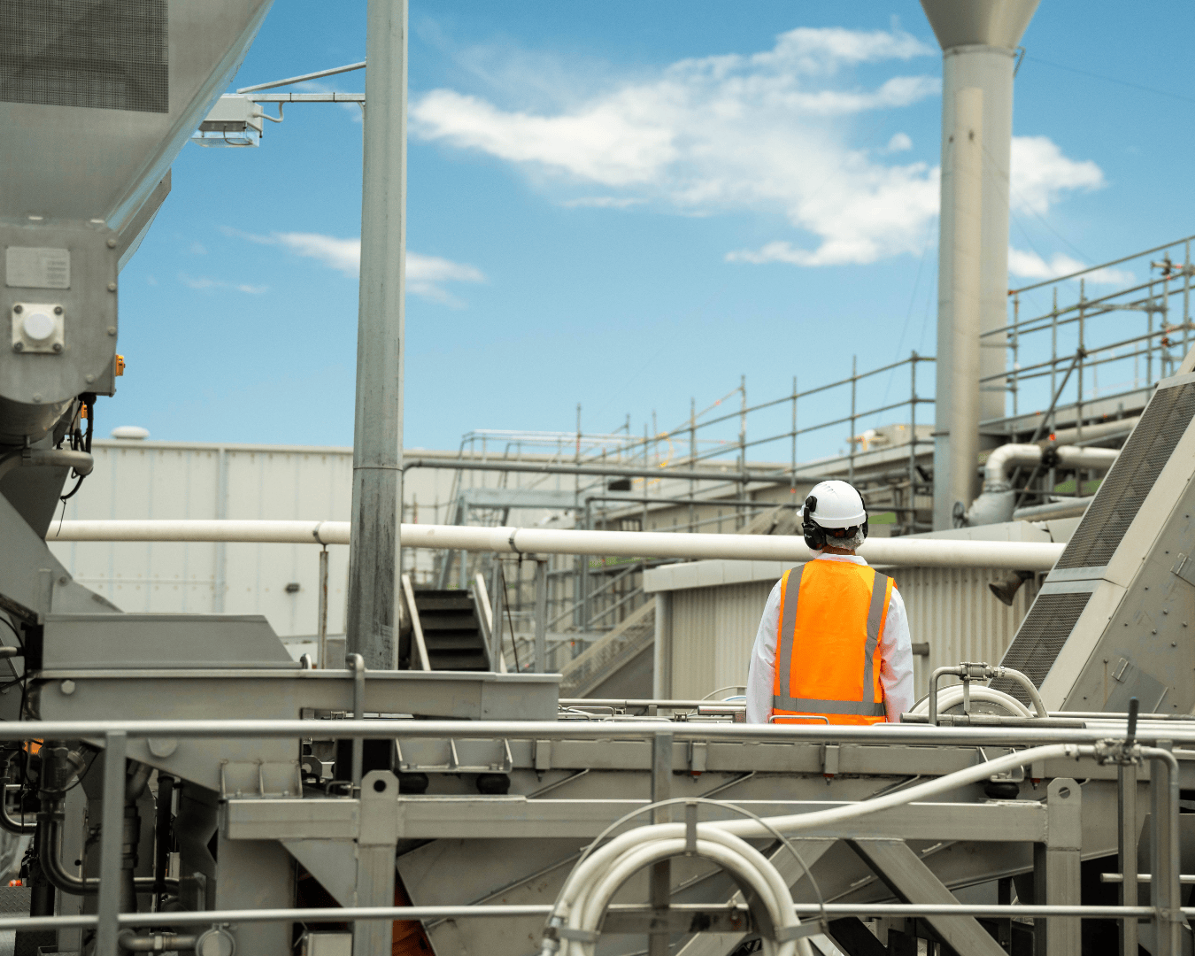 A person in a hard hat and orange safety vest stands atop metal scaffolding at a McCain facility. Visualises film content for recruitment in a large-scale manufacturing environment.