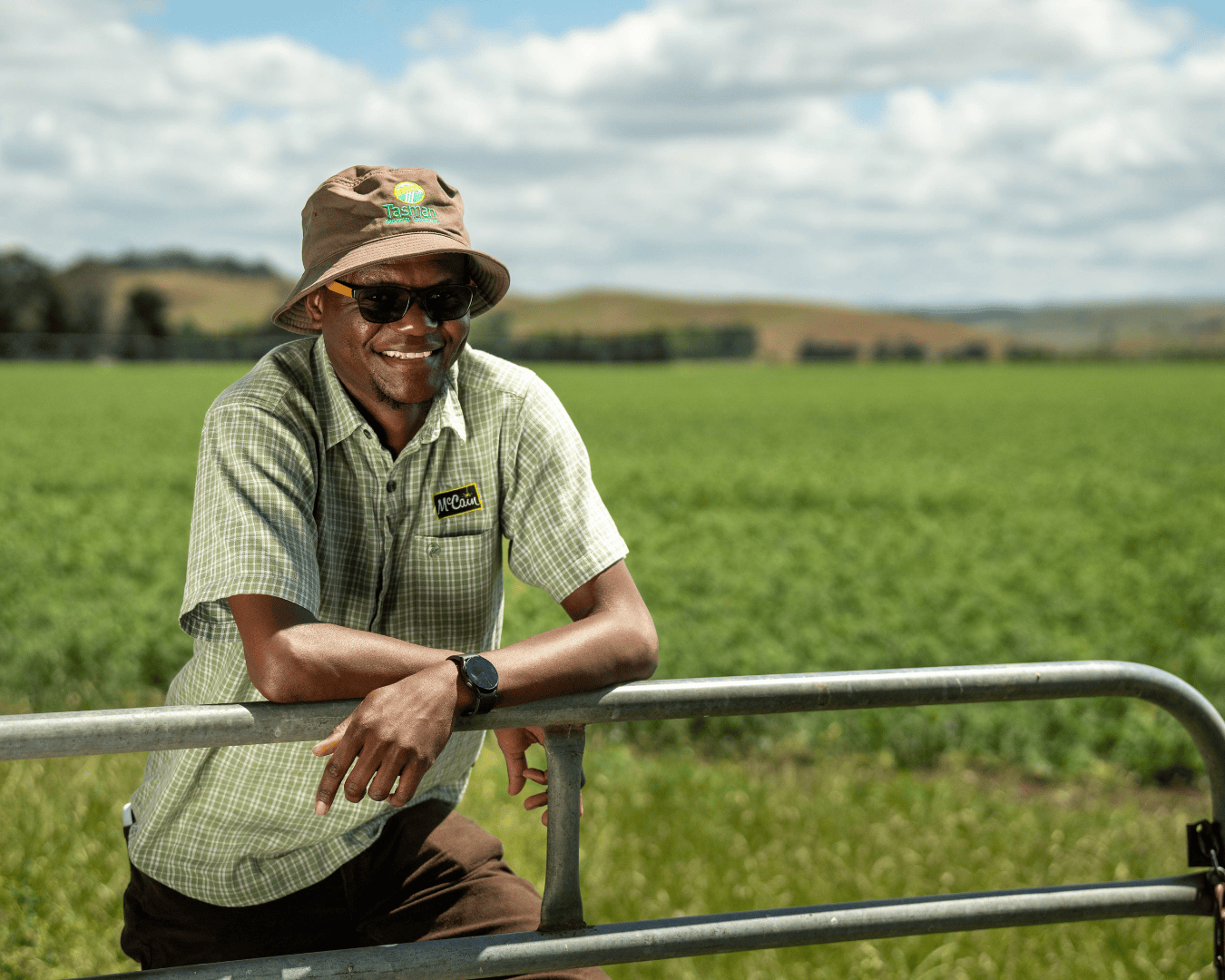 A Black man in sunglasses and a brown bucket hat smiles in front of vast green farmland. Portraying values-based employer marketing for McCain ANZ’s film-led talent attraction campaign.