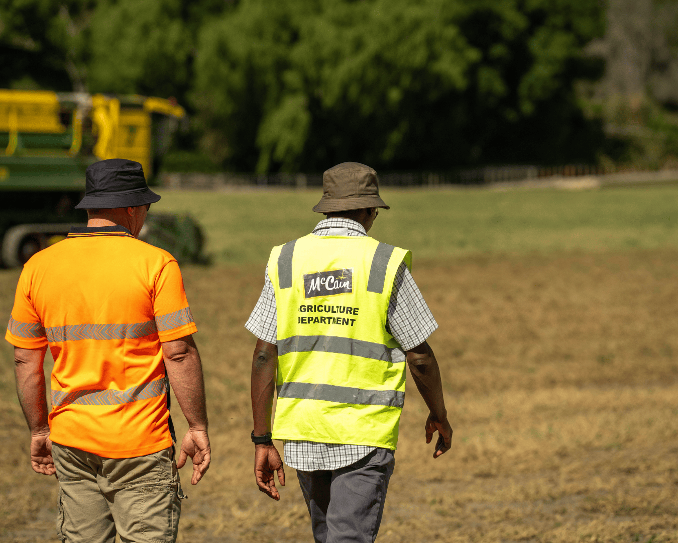 Two men walk through a dry crop field; one in an orange hi-vis shirt, the other in a McCain agriculture vest. Captures what are you made of campaign in action.