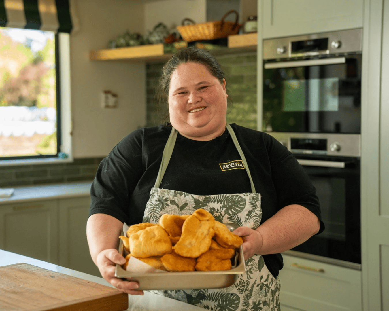 Smiling woman in a kitchen holding a tray of freshly cooked golden fry bread, wearing a McCain apron, standing in front of a modern oven.