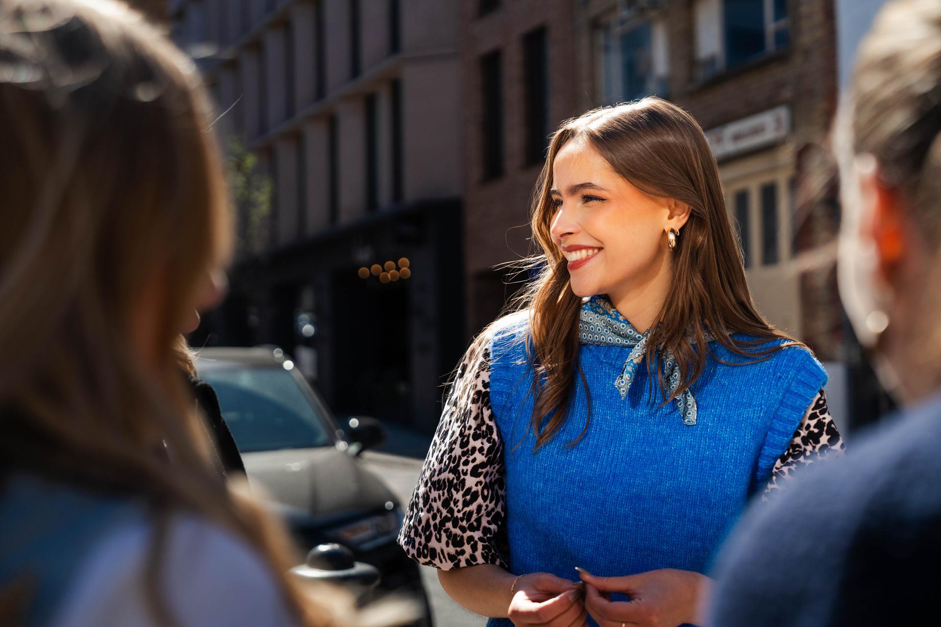 Smiling woman wearing a blue sweater, patterned scarf, and gold earrings engages in conversation with two friends on a sunny urban street lined with buildings, cars, and soft afternoon light.