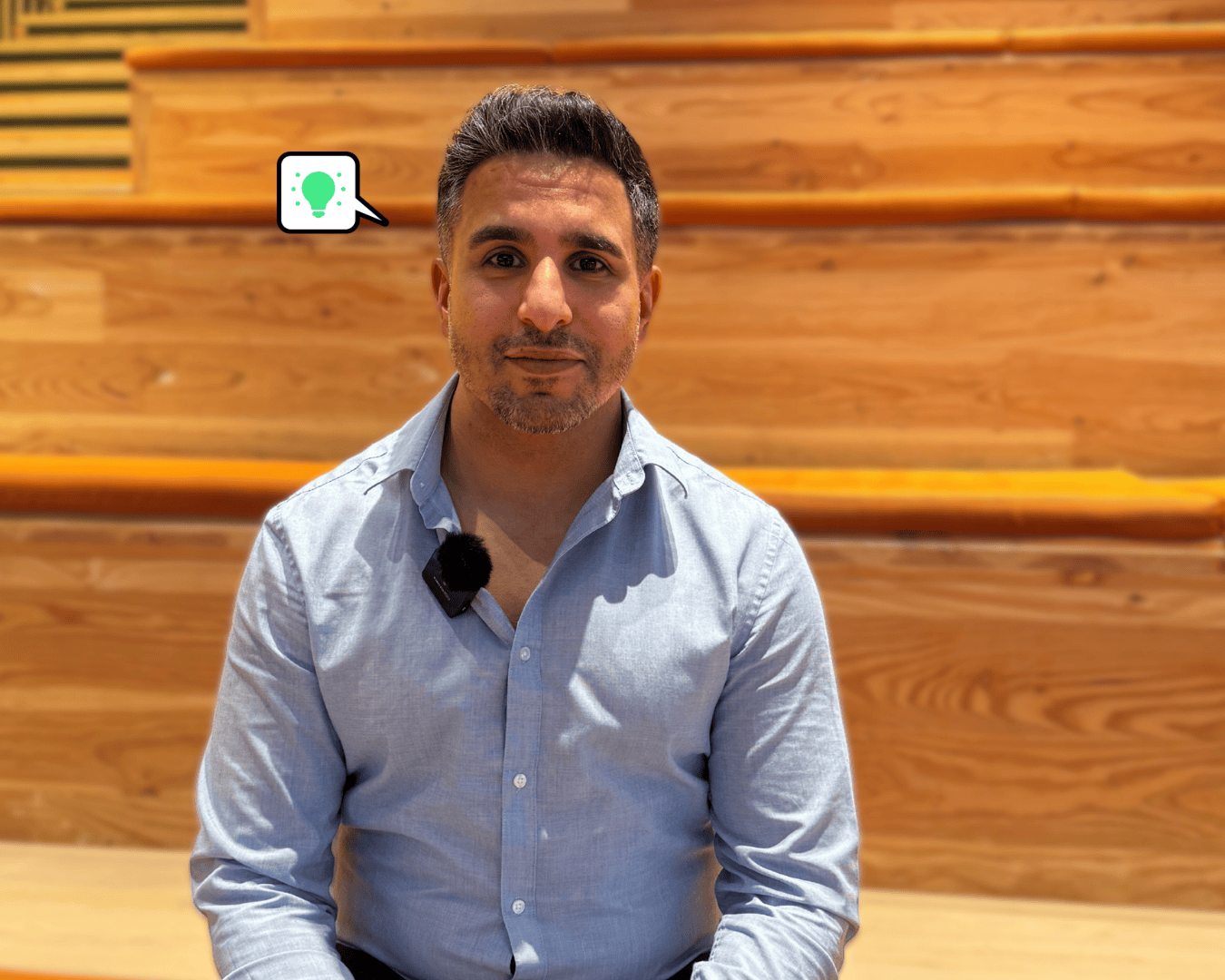 Man in light blue shirt with lapel mic, seated on tiered wooden steps, recording video content for a recruitment marketing campaign.