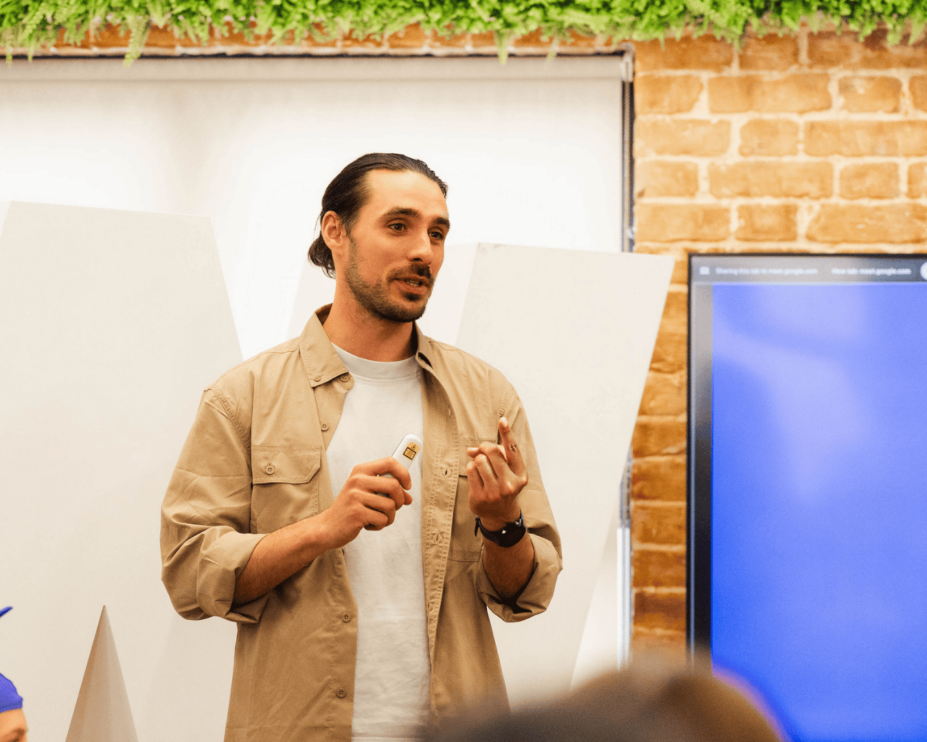 A dark-haired man with tied-back hair and light shirt presents confidently during an early talent development workshop, holding a presentation remote and facilitating learning for future professionals in a modern workspace.