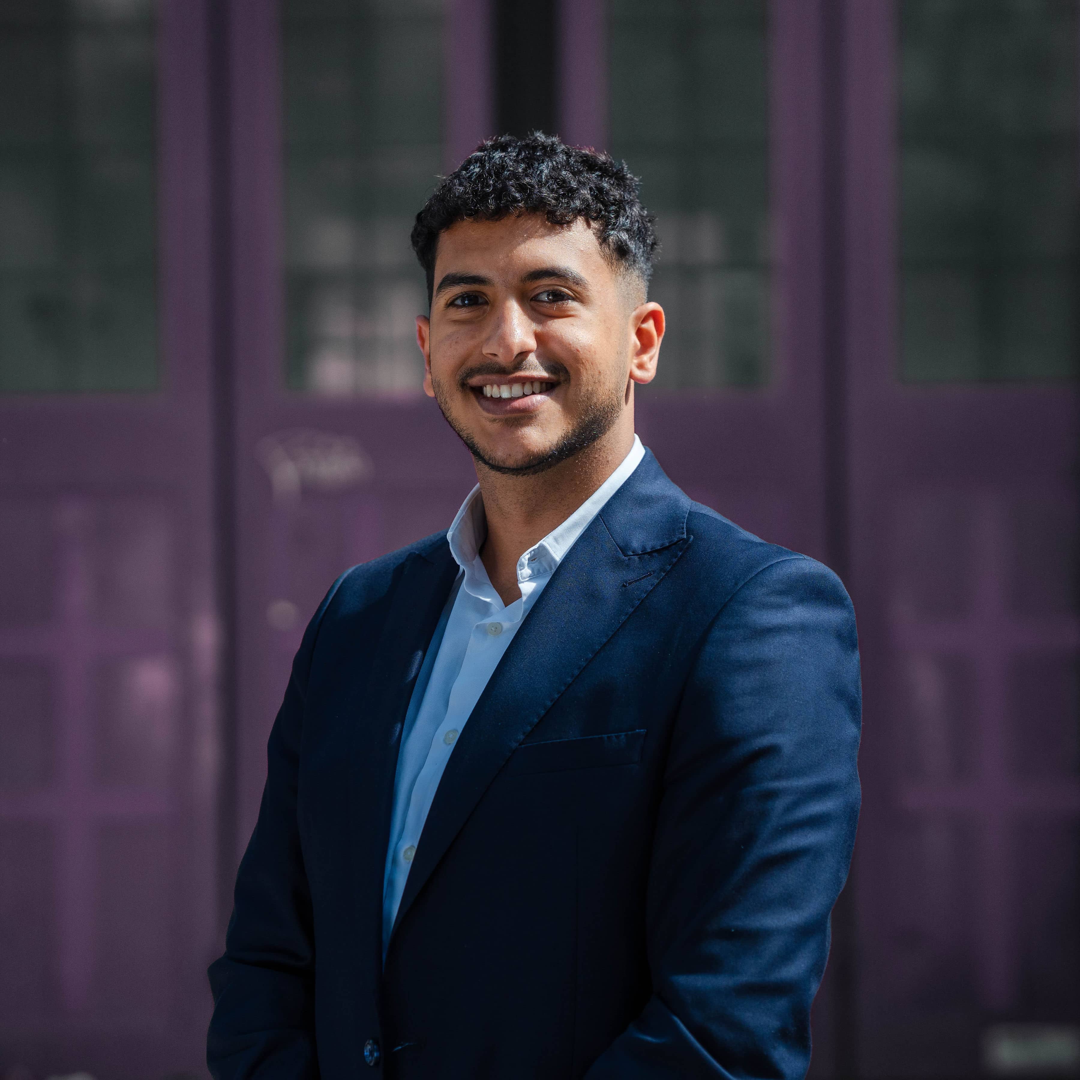 Young Middle Eastern or South Asian male graduate with short curly hair wearing a navy blue suit smiles confidently outside during early careers development programme photoshoot at Wiser Academy.