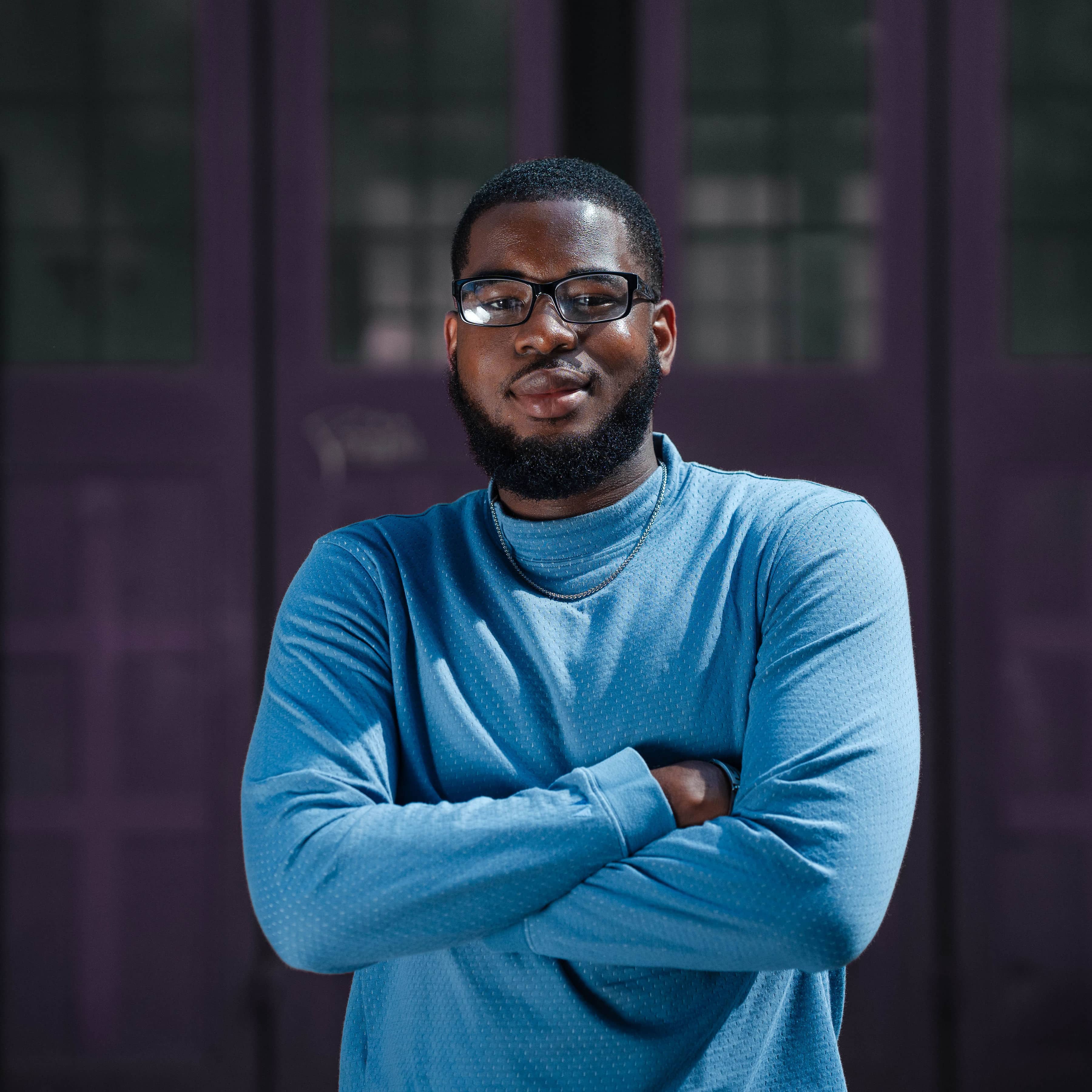 Black male graduate in blue jumper with beard and glasses stands outside with arms crossed, posing for Wiser Academy’s early careers ambassador training and professional development series.