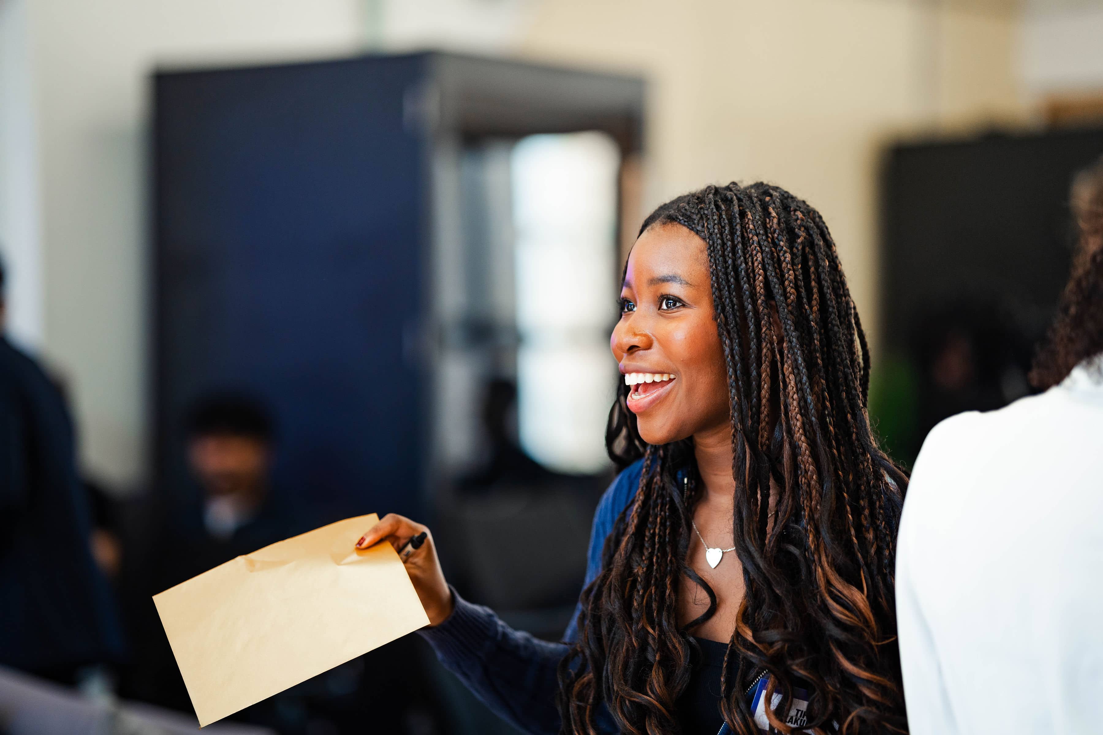 Black female graduate with long braids and navy cardigan smiles brightly while holding tan envelope during ambassador development workshop indoors.
