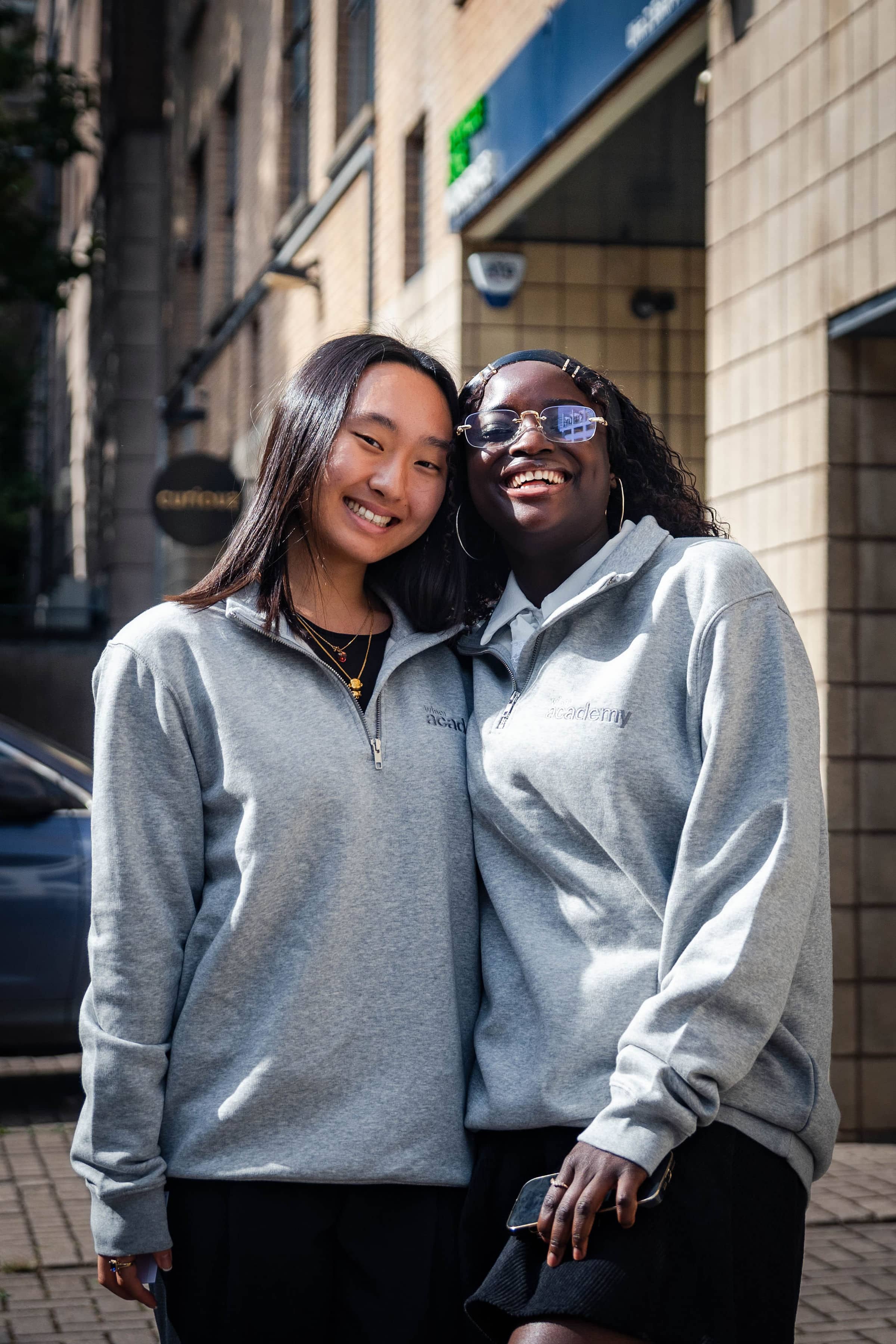 Two diverse young women, one East Asian and one Black, wearing grey Wiser Academy sweatshirts, smiling and standing outside during graduate ambassador programme.