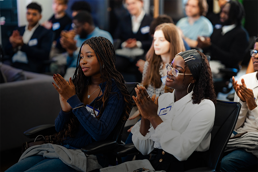 Two young women in smart casual attire clap enthusiastically at an indoor conference, surrounded by a diverse audience of similarly aged adults seated in rows, all engaged and focused on the presentation.
