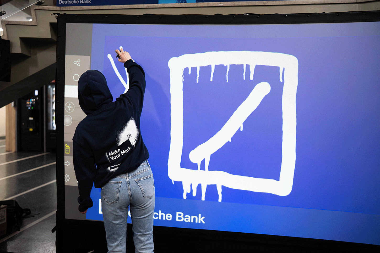 Back view of a person in a navy hoodie marked “Make Your Mark” painting a stylised Deutsche Bank logo on a large interactive blue screen inside a modern building interior.