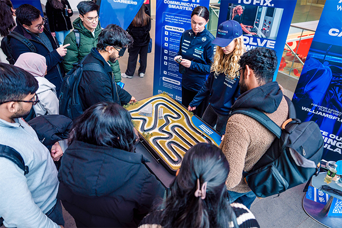 Large group of students gathers at a careers event, engaging with a tabletop racing circuit run by Williams team members in branded outfits, surrounded by banners promoting STEM opportunities and innovation.