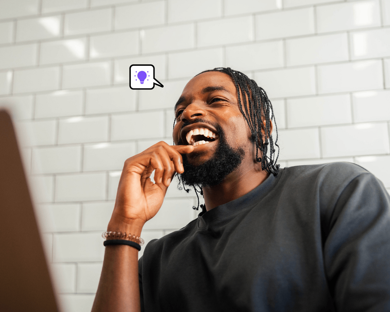Youn man in black tshirt smiling while working on a laptop, sitting against a white tiled wall with a purple idea icon