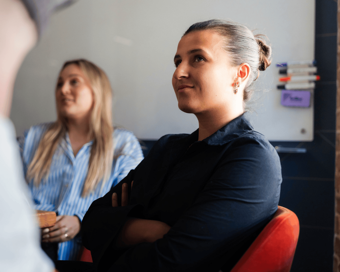 Two women sit in a meeting room, engaged in conversation. The woman in the foreground wears a dark shirt and has her arms folded, looking ahead thoughtfully. The second woman, slightly out of focus in the background, wears a blue and white striped shirt and appears to be speaking. A whiteboard with colourful markers is visible behind them.