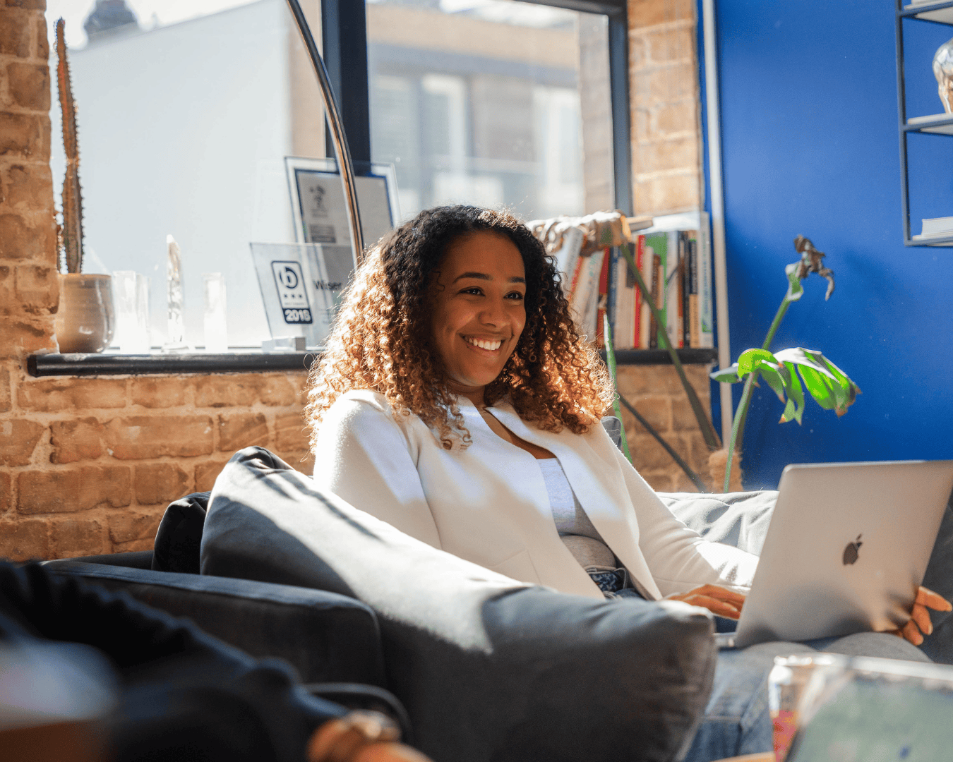 A woman sits on a sofa in a bright, modern office space, smiling while working on a laptop. She’s wearing a white blazer and blue jeans, with sunlight streaming through large windows behind her. A brick wall, bookshelves, and a framed award add warmth and character to the background.