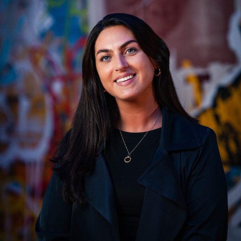 Portrait of a smiling woman with long dark hair and gold hoop earrings, wearing a black top and necklace, posing confidently in front of a colourful, urban-style painted wall background.