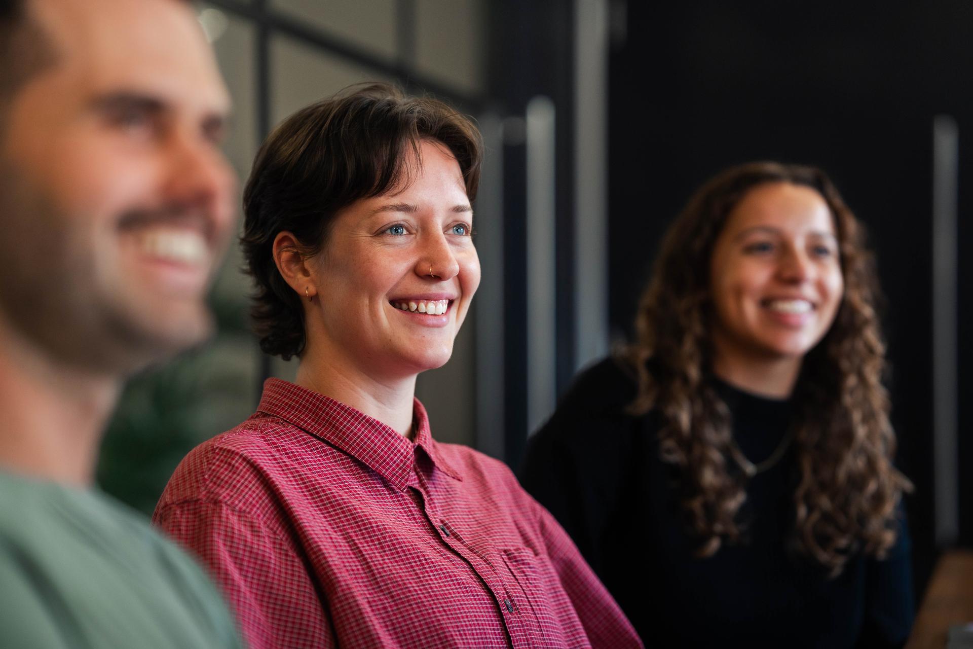 A group of three smiling coworkers attentively listen during an informal team meeting in a modern office setting, with natural light and glass partitions creating a welcoming professional atmosphere.
