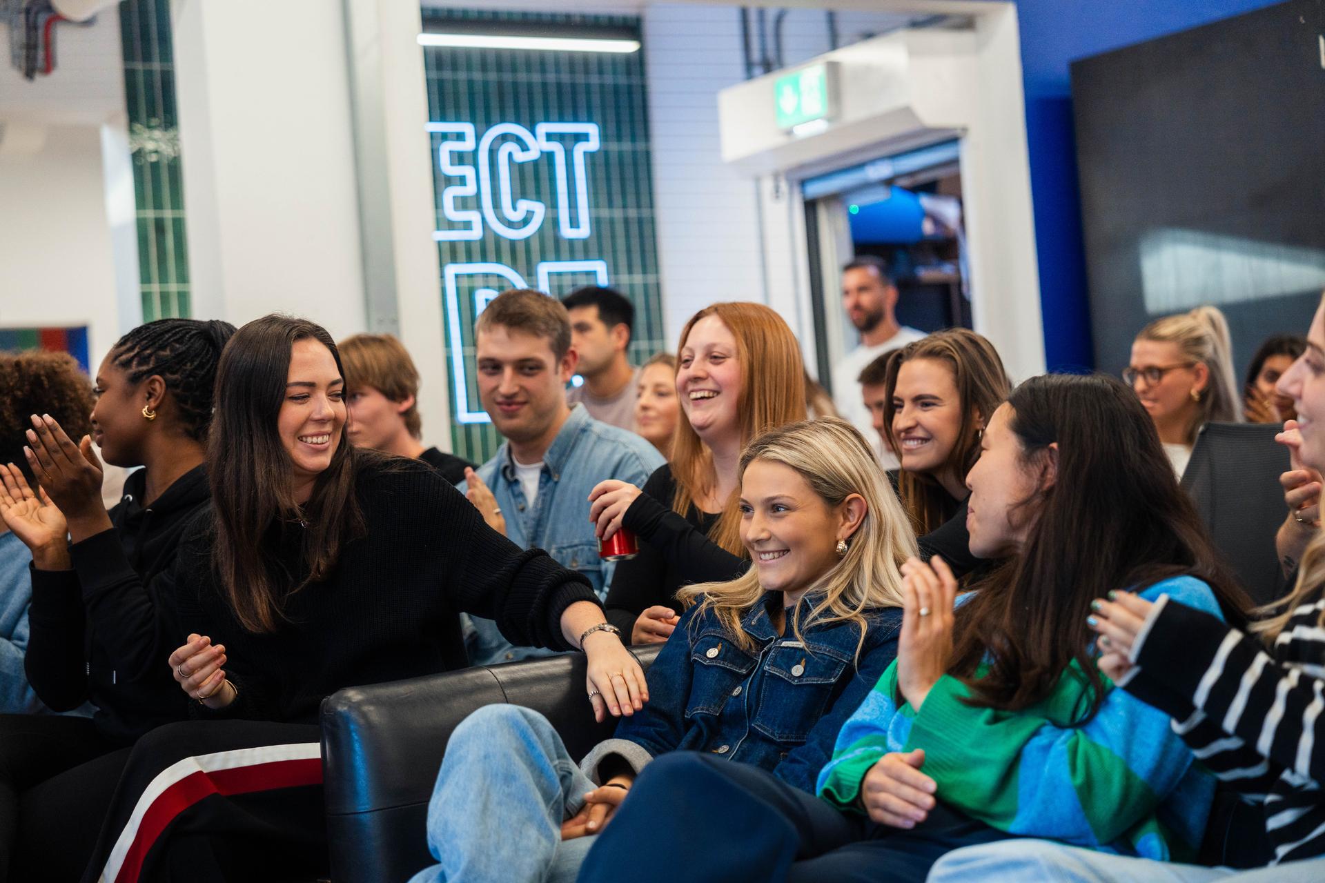 A diverse team of coworkers smile, laugh, and applaud during a relaxed office event, surrounded by modern design elements like neon signage, open seating, and a collaborative atmosphere.