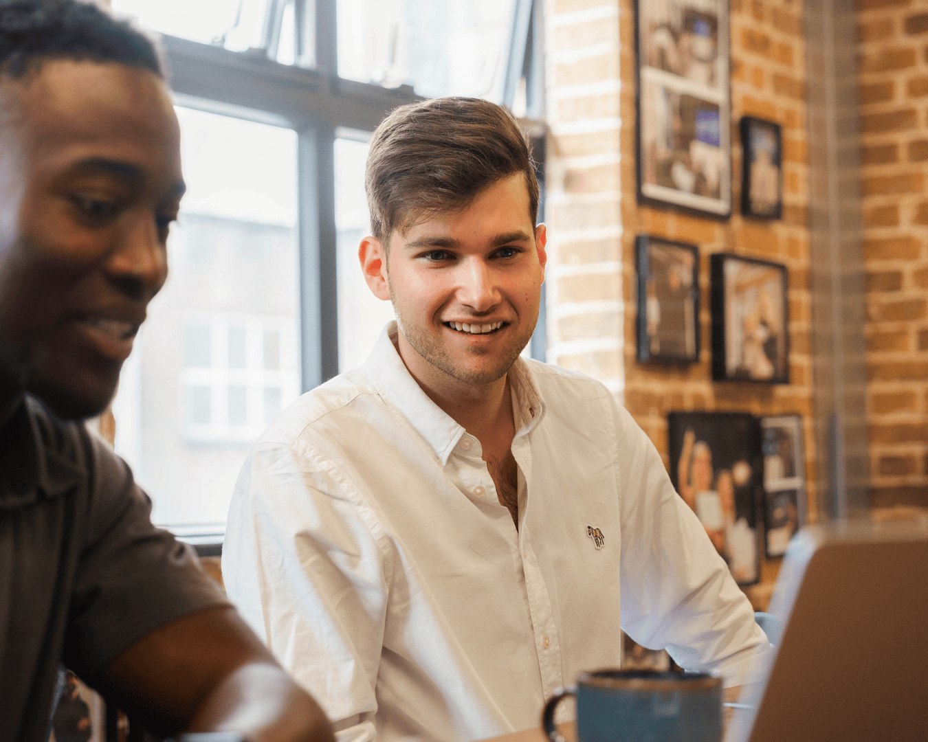 Two professionals working together in a casual office setting. One man, smiling in a white shirt, looks toward the camera while seated at a laptop. Brick walls and framed photos create a warm, collaborative backdrop.