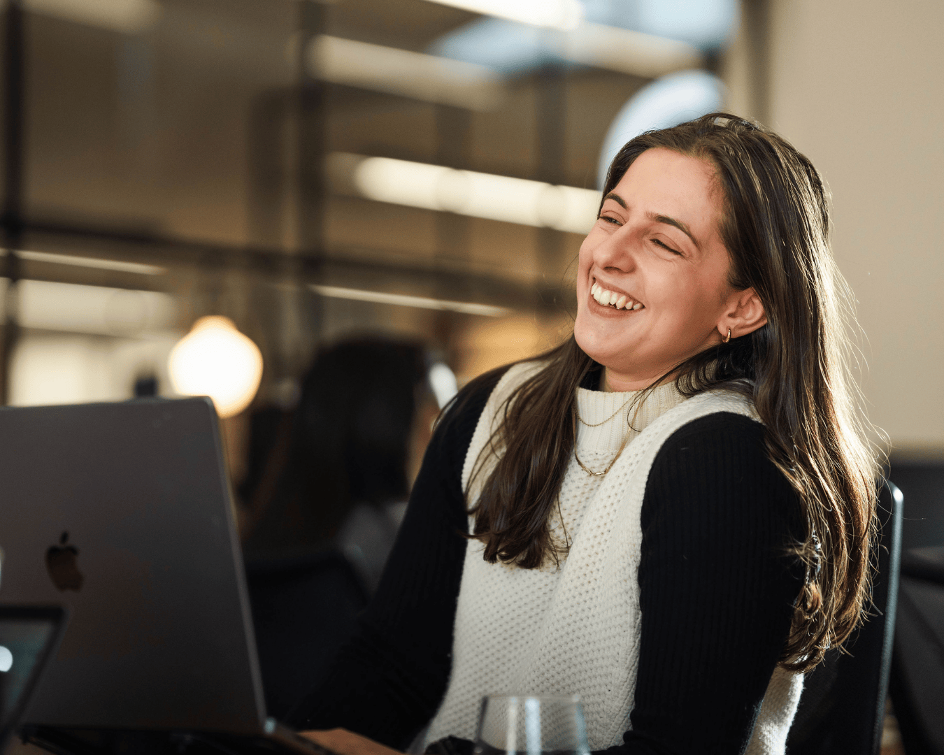 A candid photo of a woman mid-laugh at her desk, lit by warm natural light. She’s surrounded by the soft blur of a modern office, with a laptop open in front of her.